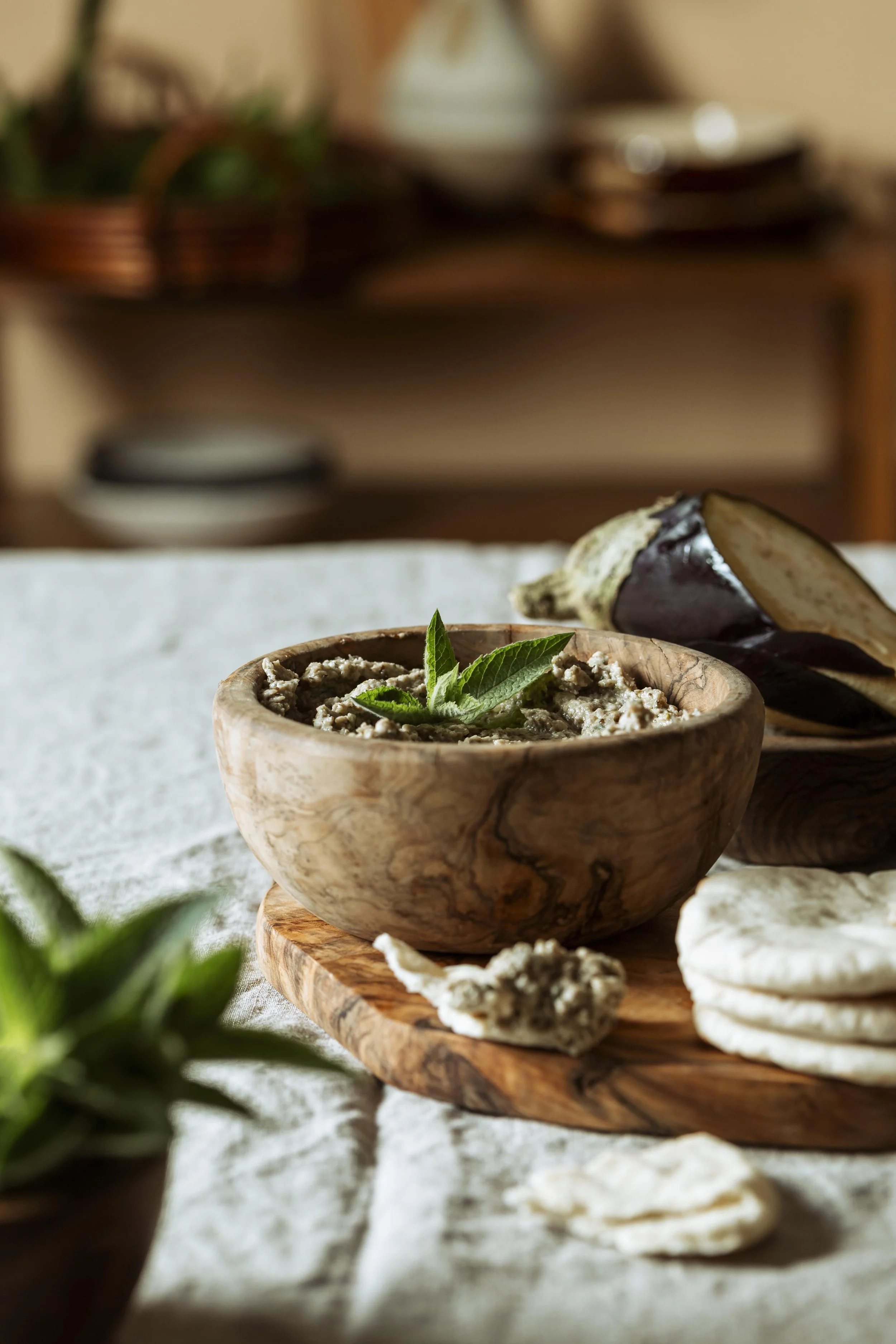 Close-up of a bowl of babaganoush garnished with a green herb leaf, on a rustic wooden serving board, with eggplant and flatbread nearby, set on a textured cloth surface.