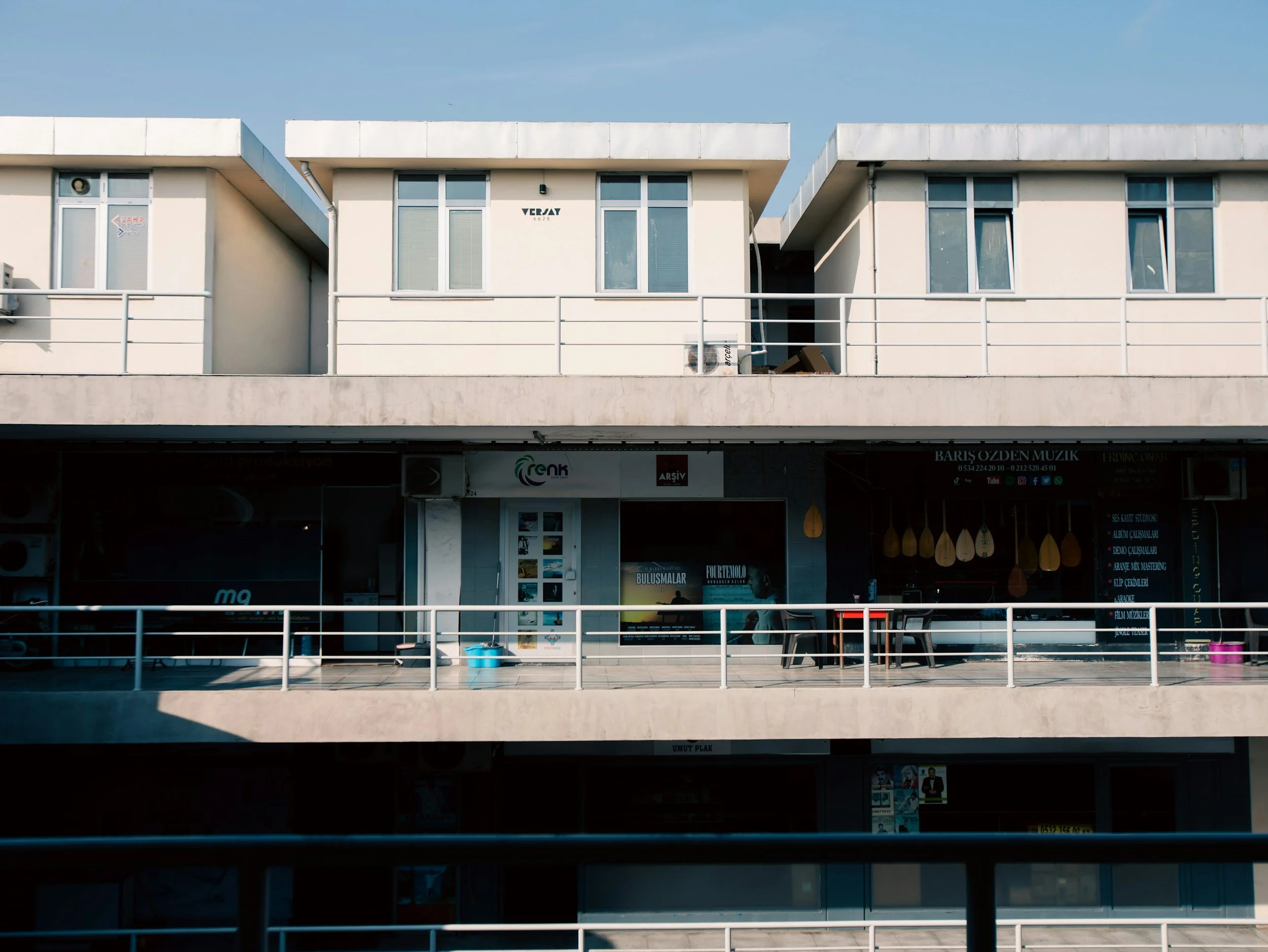 Three-story beige commercial building with storefronts and windows, and a small balcony with a white railing in front. Clear blue sky overhead.