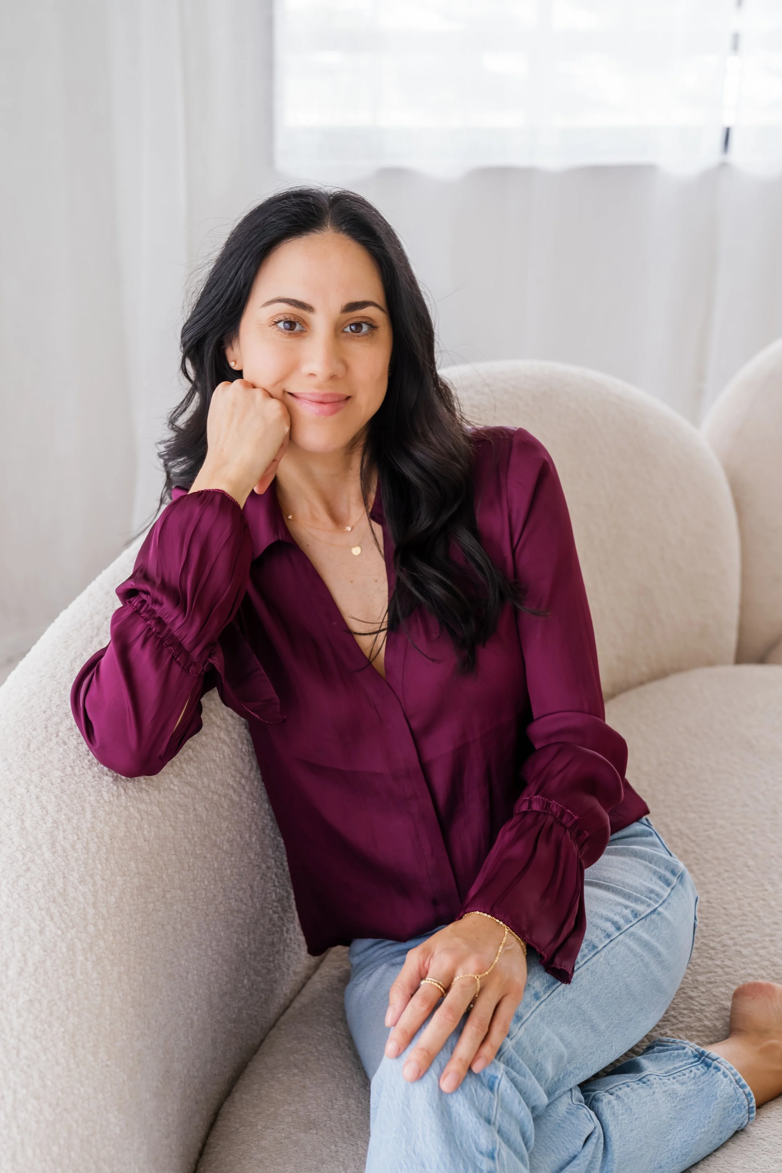 A woman with long dark hair and light skin is sitting on a beige couch, resting her chin on her hand and smiling at the camera. She is wearing a burgundy blouse and light blue jeans, with a gold bracelet and necklace, in a bright, minimalistic room with sheer white curtains.