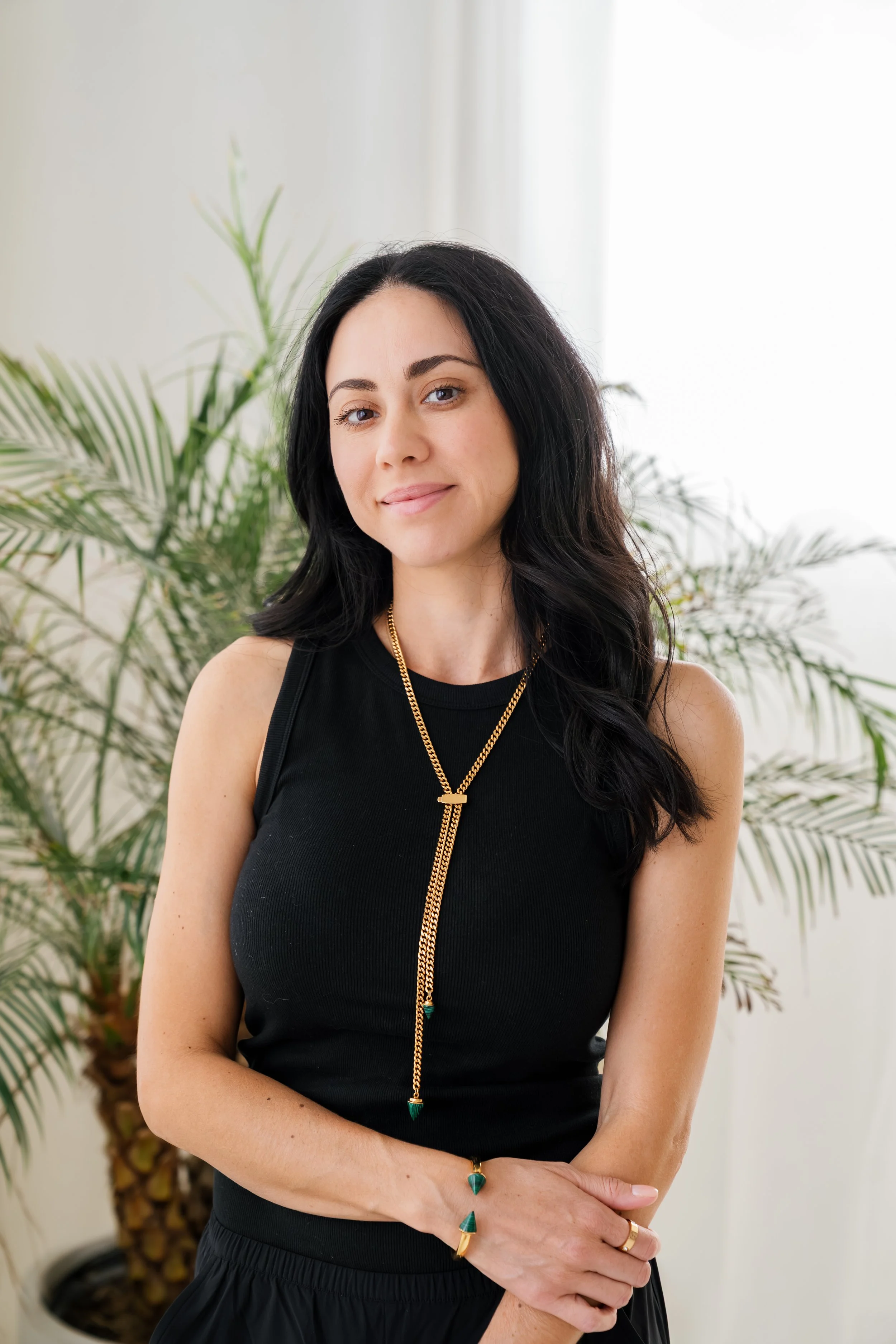 A woman with long black hair and light skin, wearing a sleeveless black top with gold jewelry, standing indoors in front of a leafy green plant with a blurred light-colored background.