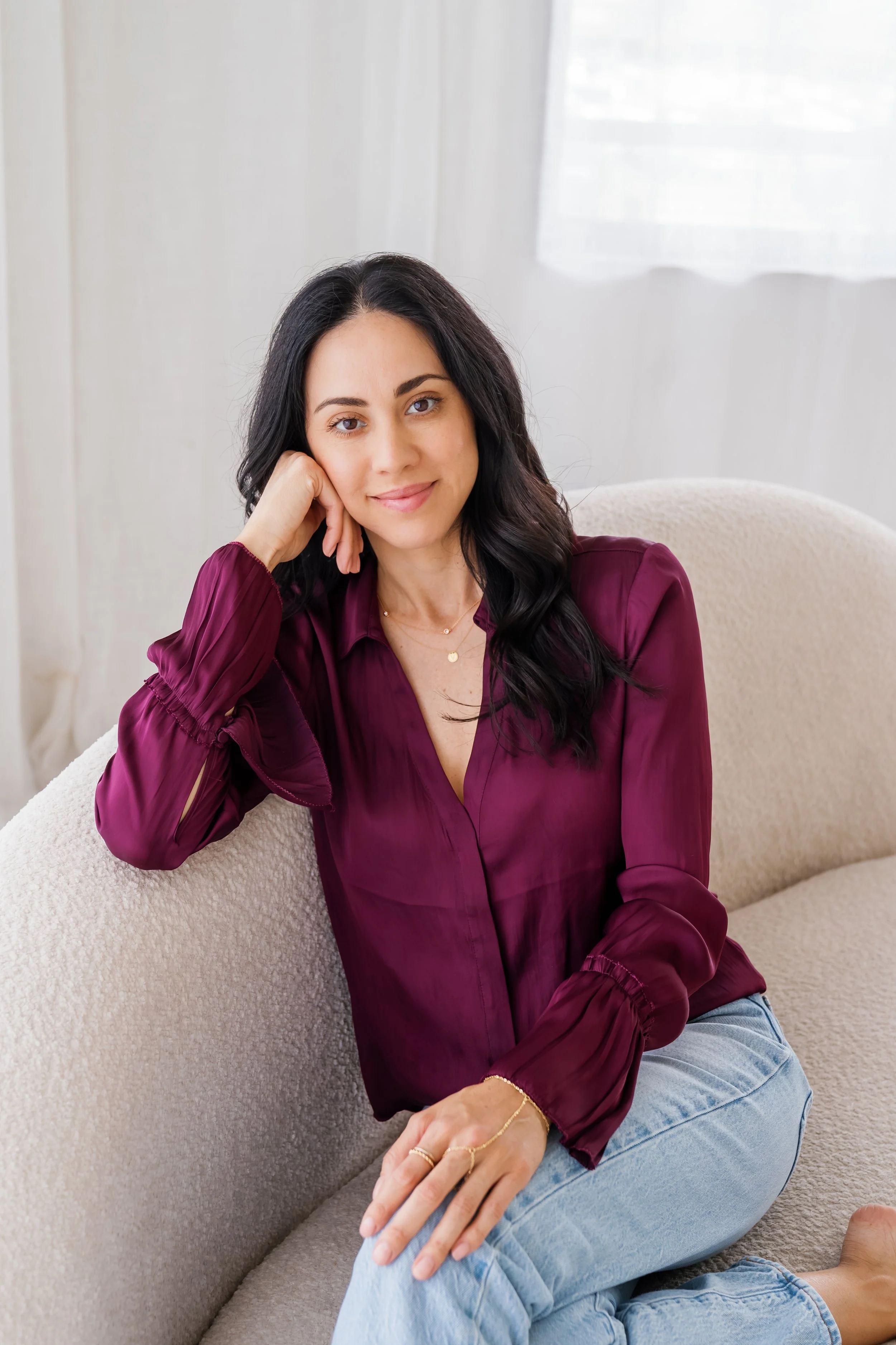 A woman with black hair, wearing a burgundy silky blouse and light blue jeans, sitting on a beige couch. She is resting her head on her left hand, smiling softly at the camera, with a sheer white curtain behind her.