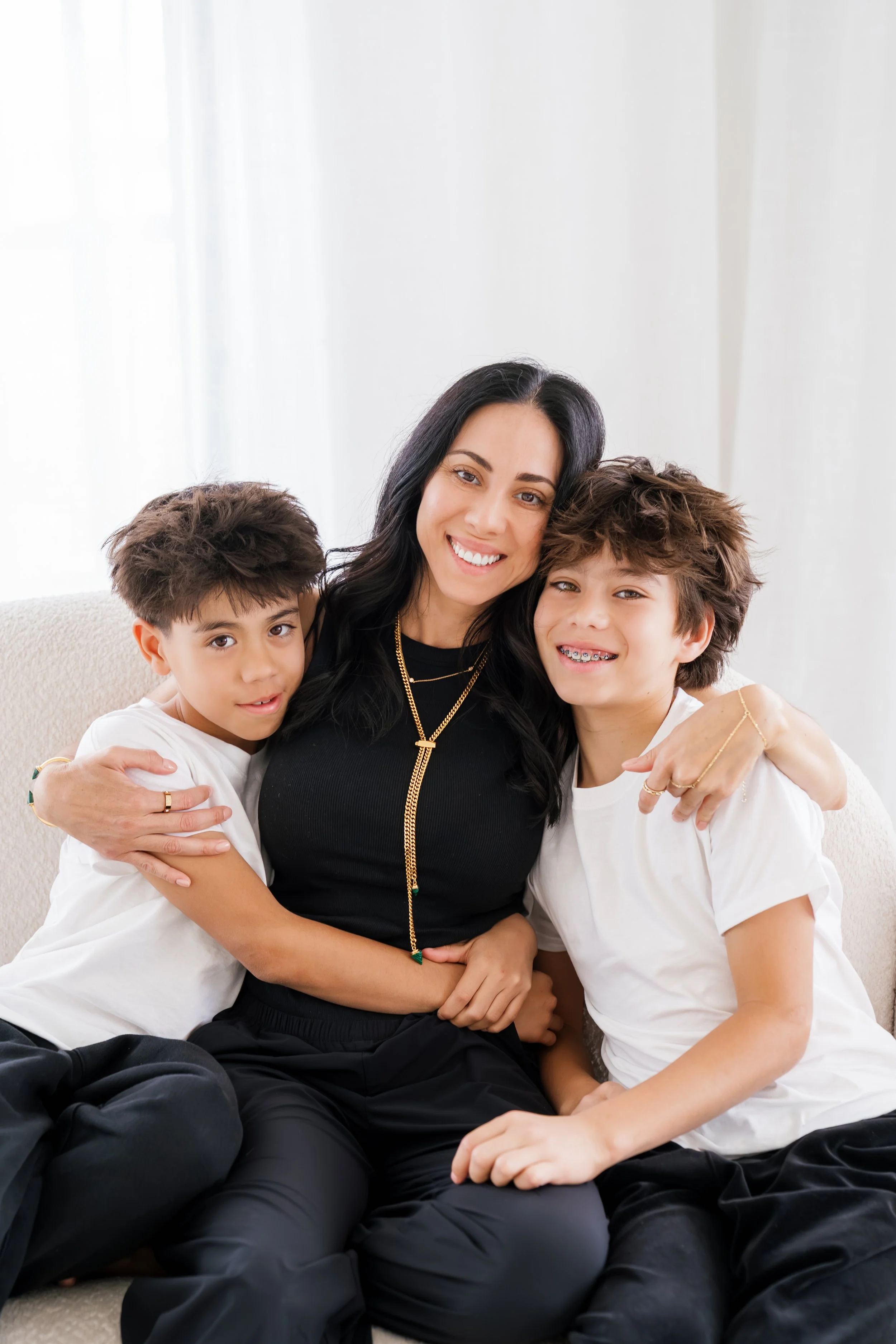A woman with long dark hair smiling and hugging two boys, all sitting on a beige couch against a white background. The boys have curly hair and are wearing white shirts, with one smiling showing braces and the other with a neutral expression.