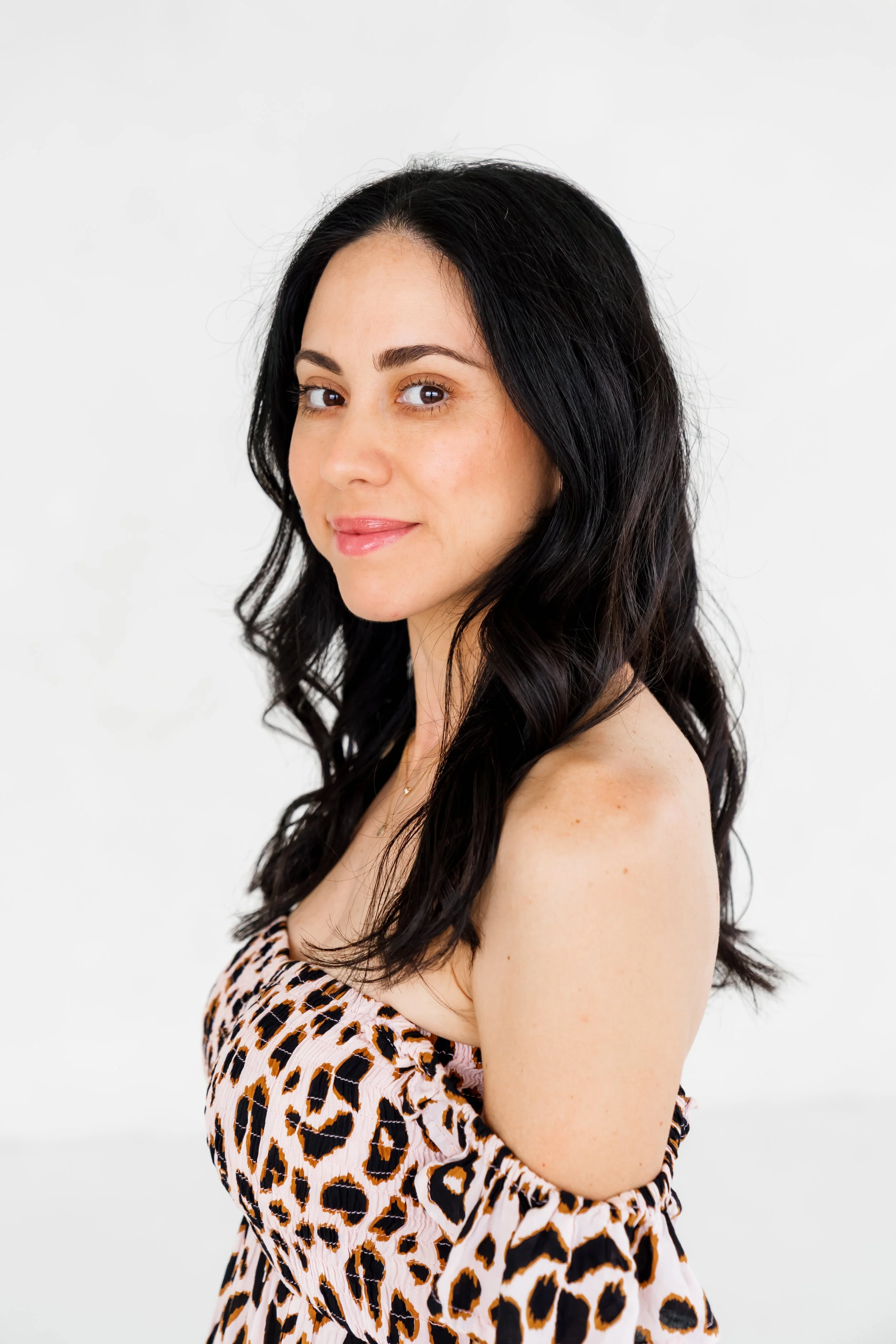 A woman with long dark hair and light skin looking over her shoulder, wearing a leopard print dress against a plain white background.