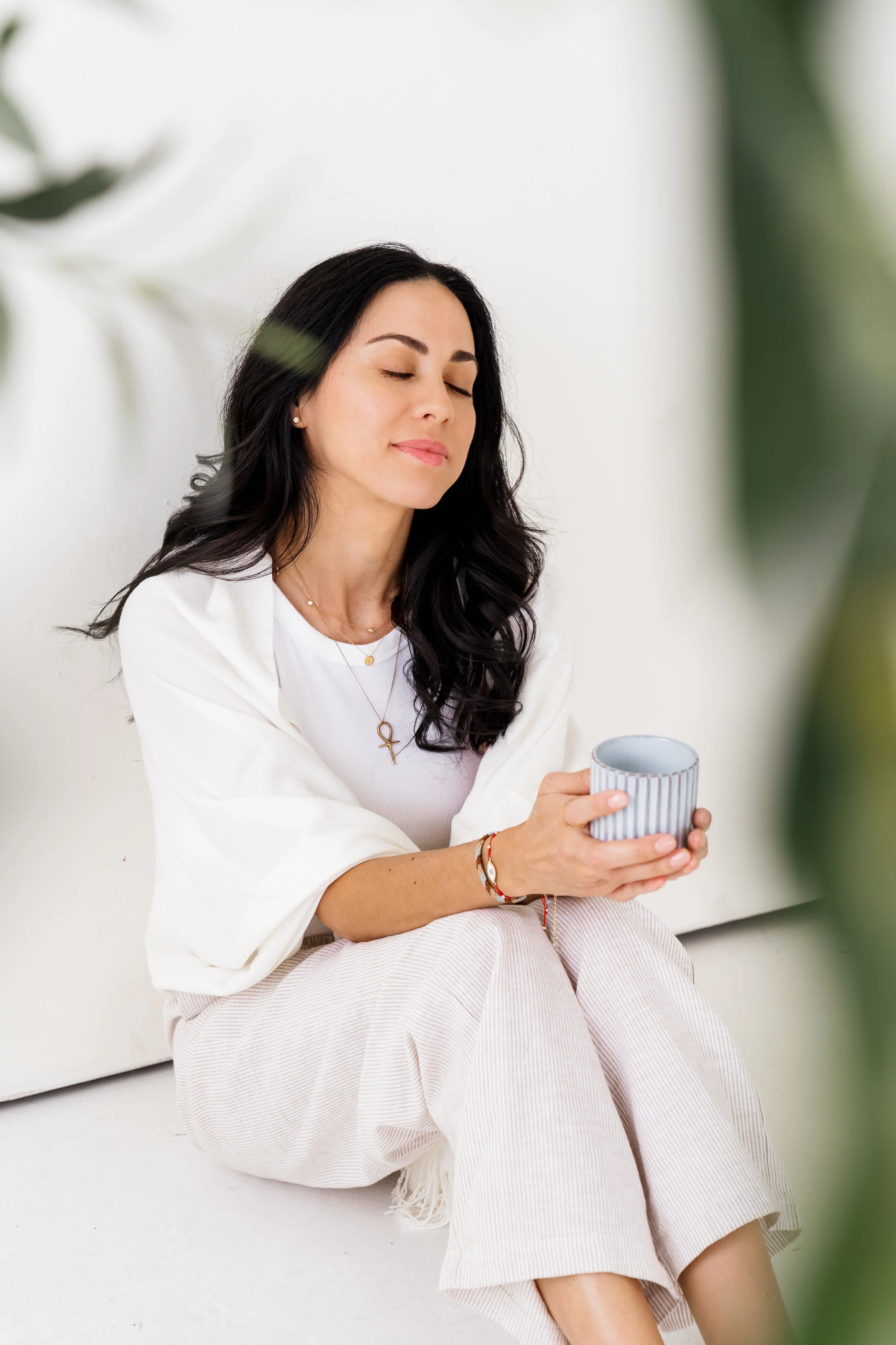 A woman with long dark hair sitting with her eyes closed, holding a striped mug, wearing a white top and beige striped pants, in a minimalistic bright room.