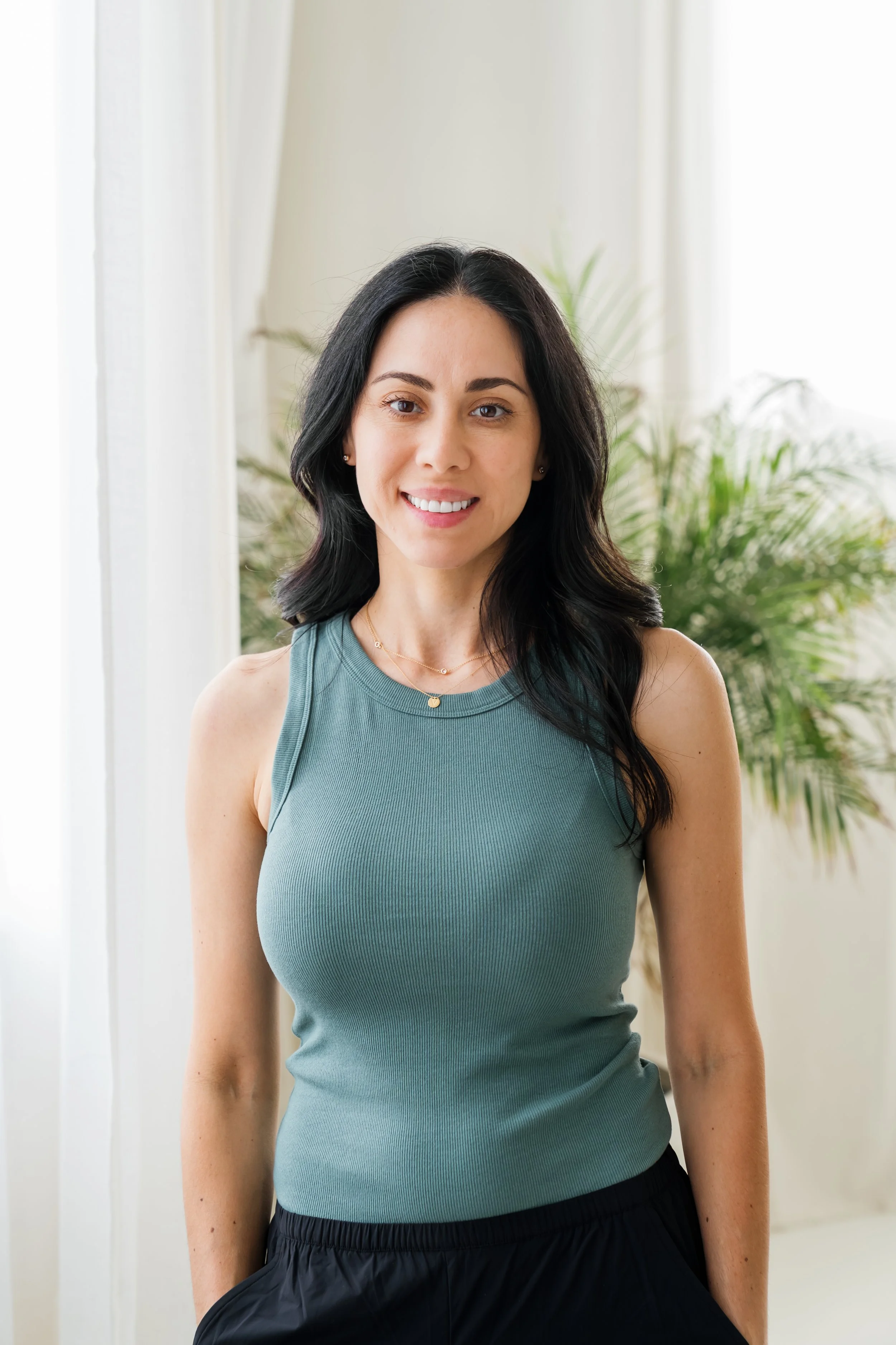 A woman smiling with black hair, wearing a teal sleeveless top and black pants, standing in front of light-colored curtains and green plants.