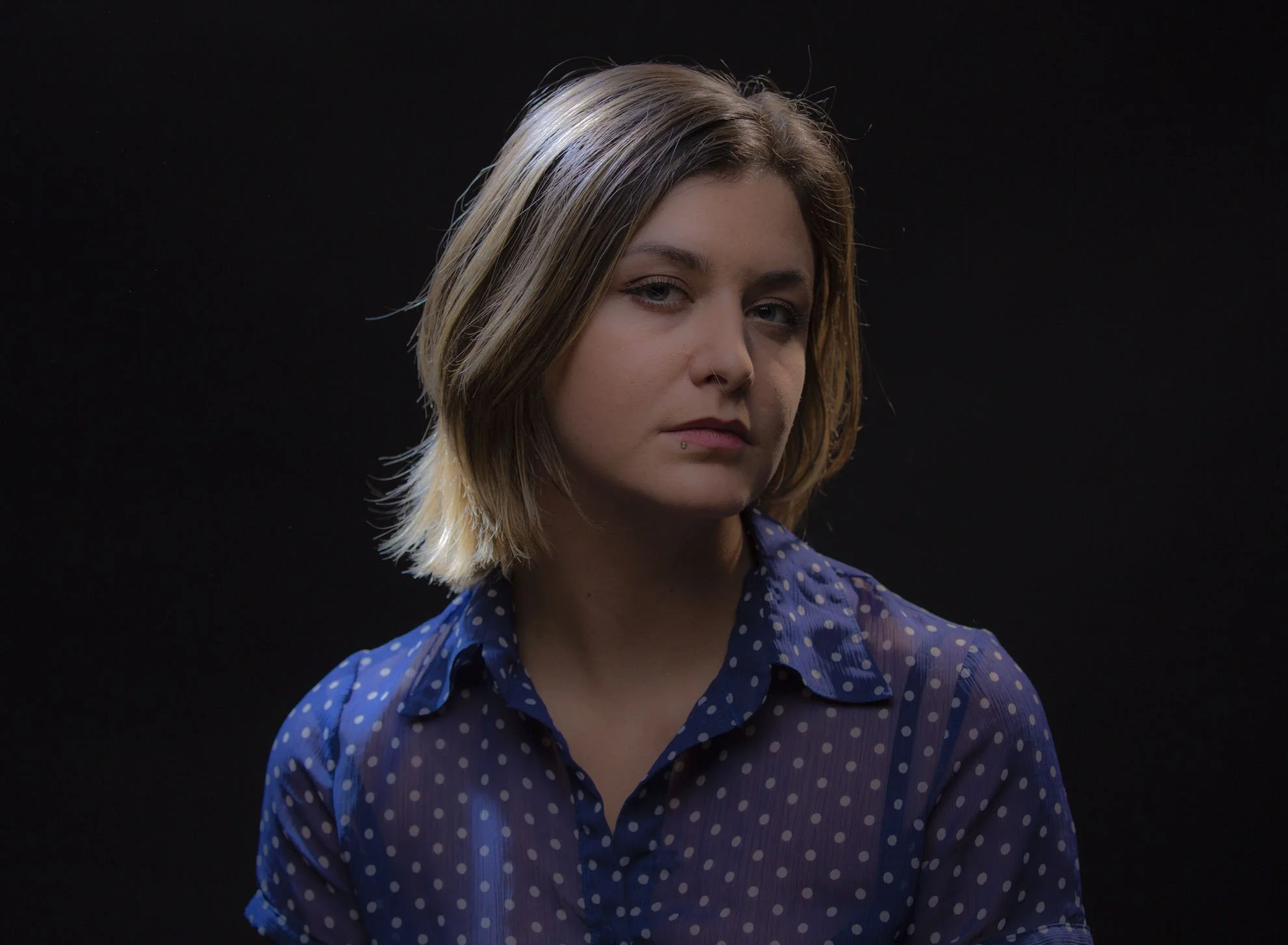 Portrait of a young woman with shoulder-length blonde hair, wearing a blue polka dot shirt, against a black background.