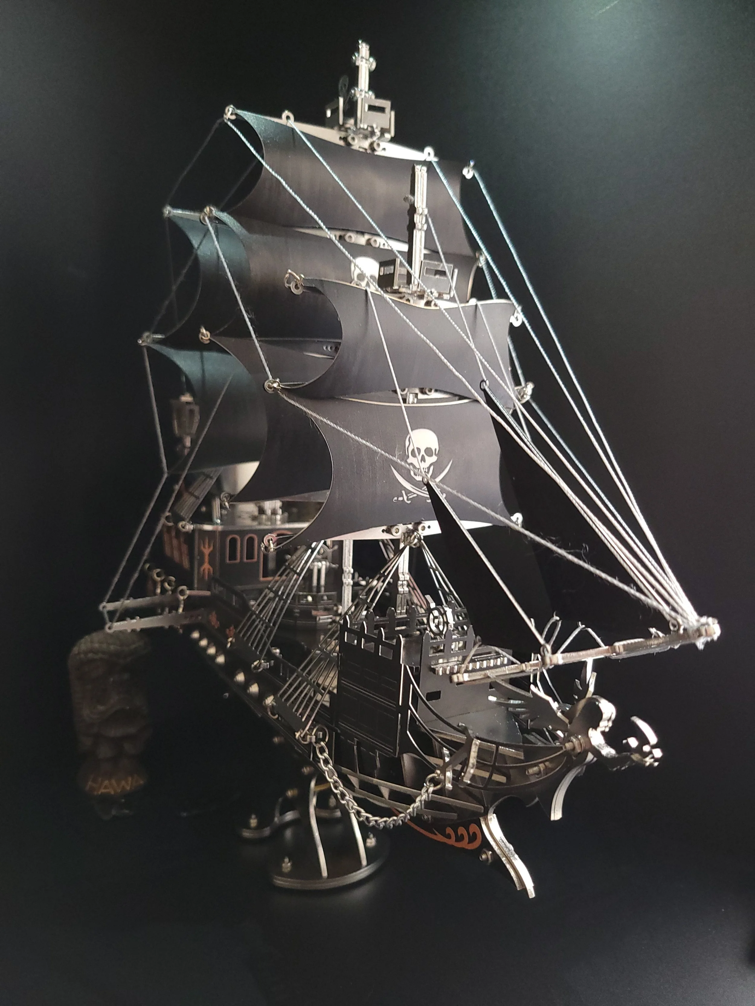 A detailed metal model of a pirate ship with black sails featuring a skull and crossbones symbol, ropes, and chain details. The ship is set against a black background.