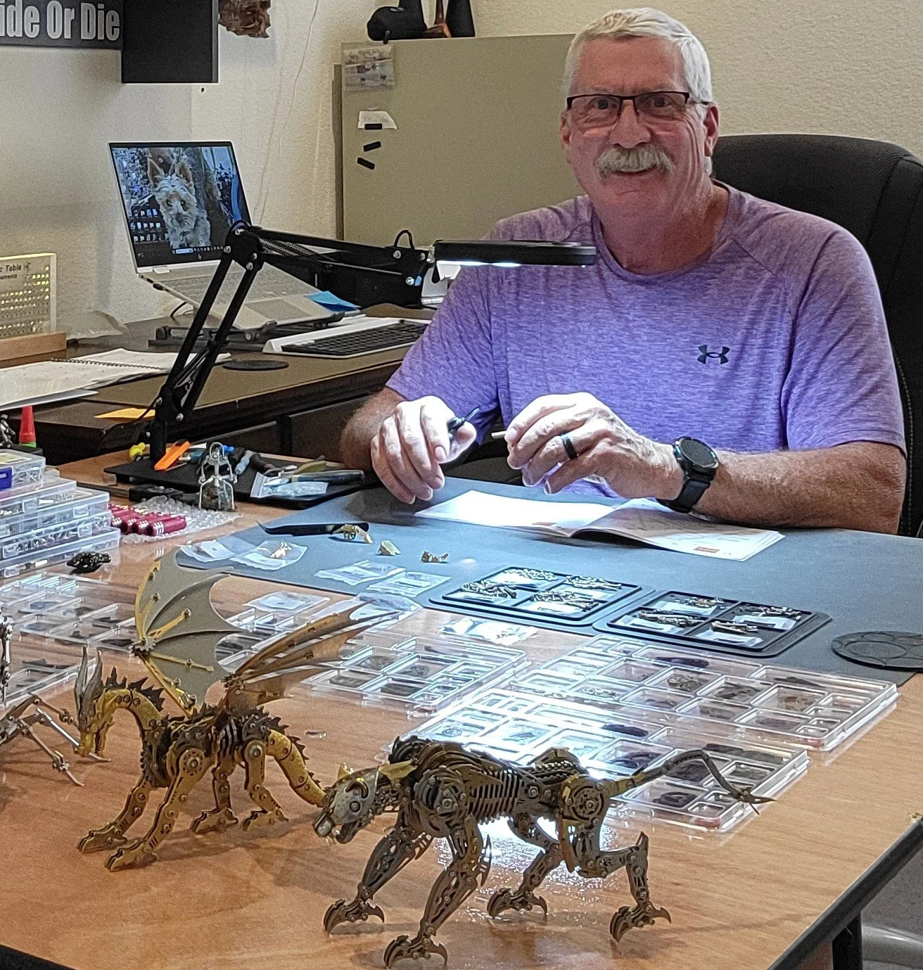 An older man with glasses and a white mustache, wearing a gray Under Armour shirt, is sitting at a desk with various miniature mechanical animal models and parts. He is working on a project with tools and papers, with a laptop showing a dog on the screen in the background.