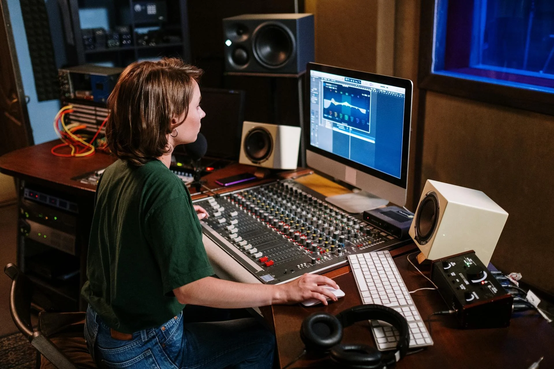 A woman in a green shirt and jeans working with sound mixing equipment in a recording studio, with computer screens and speakers around her.