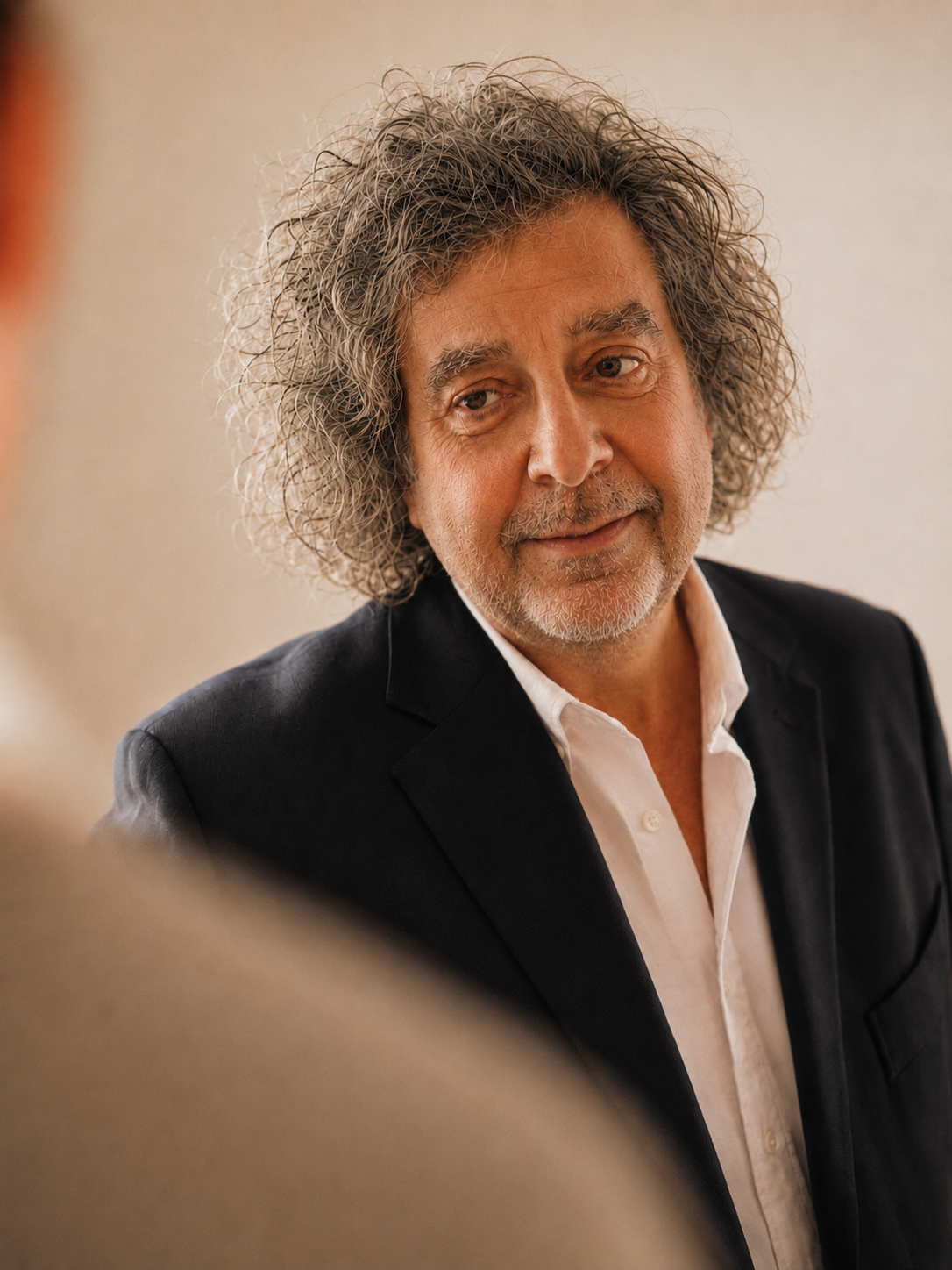 A middle-aged man with gray, curly hair and a beard, wearing a black blazer and white shirt, looking thoughtfully to the side.