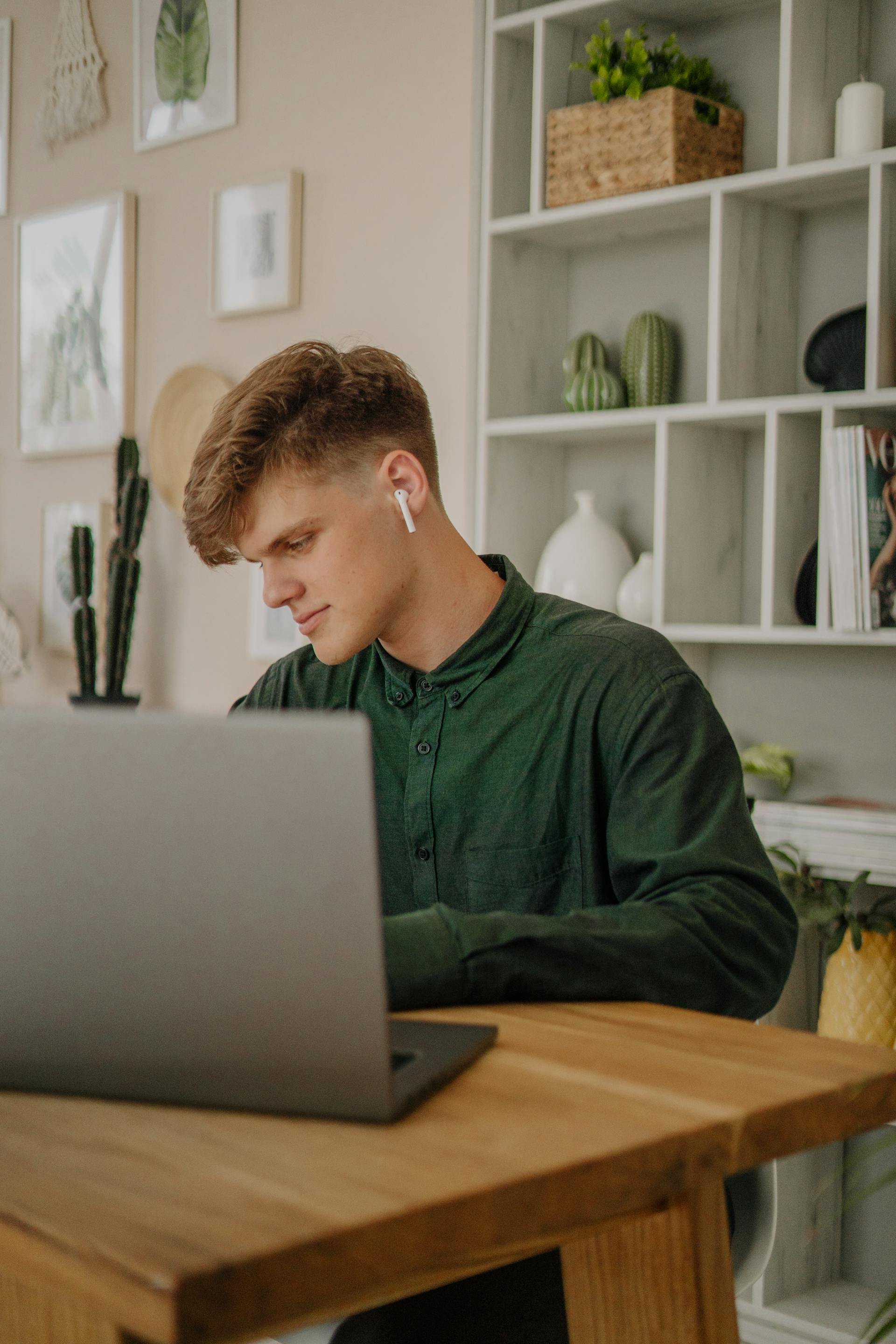 a young man sitting at a wooden desk, looking at a laptop, wearing white wireless earbuds, in a modern room with shelves and artwork on the wall.