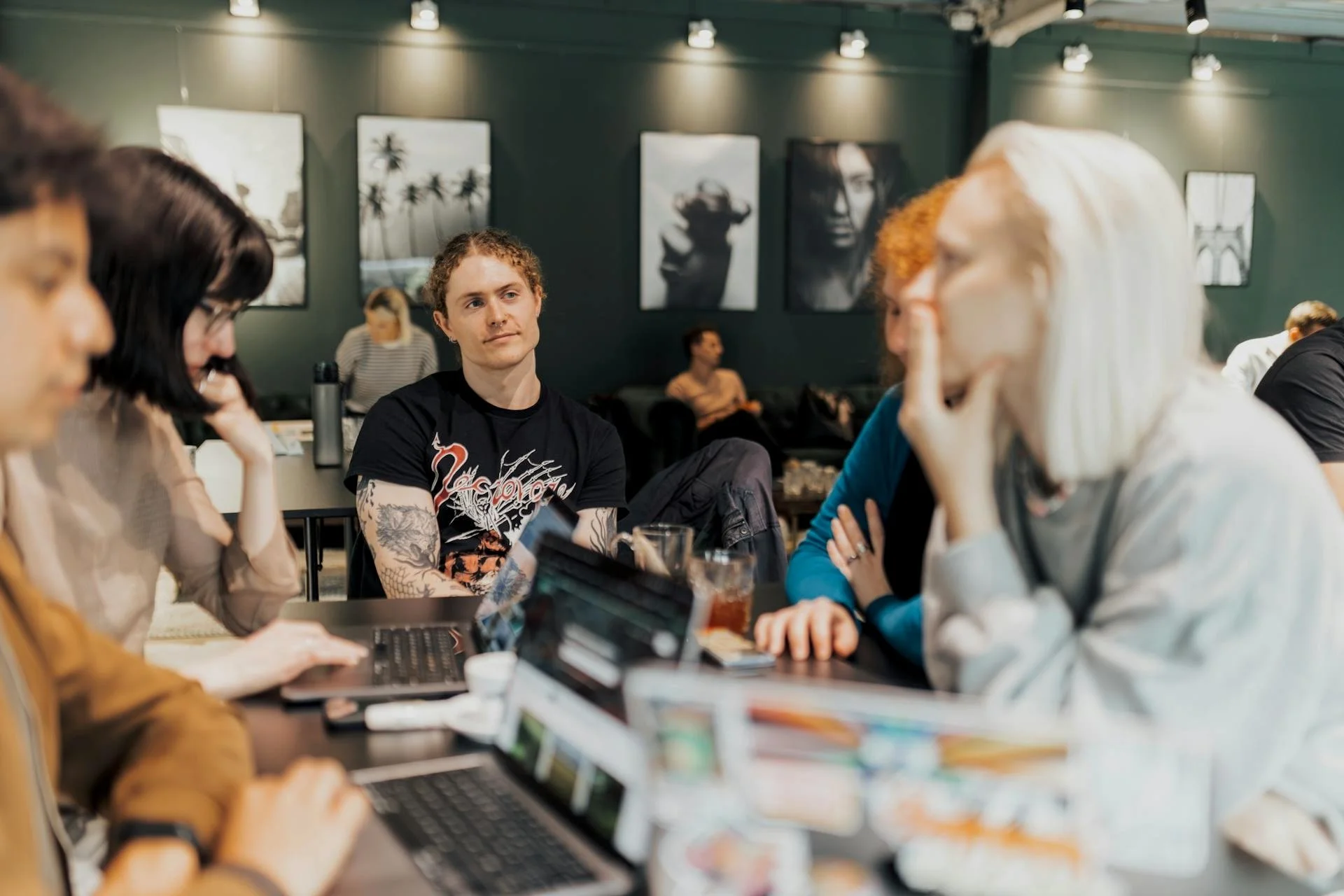 Group of people sitting at a table in a cafe or restaurant, engaged in conversation, with artwork on the dark wall behind them.