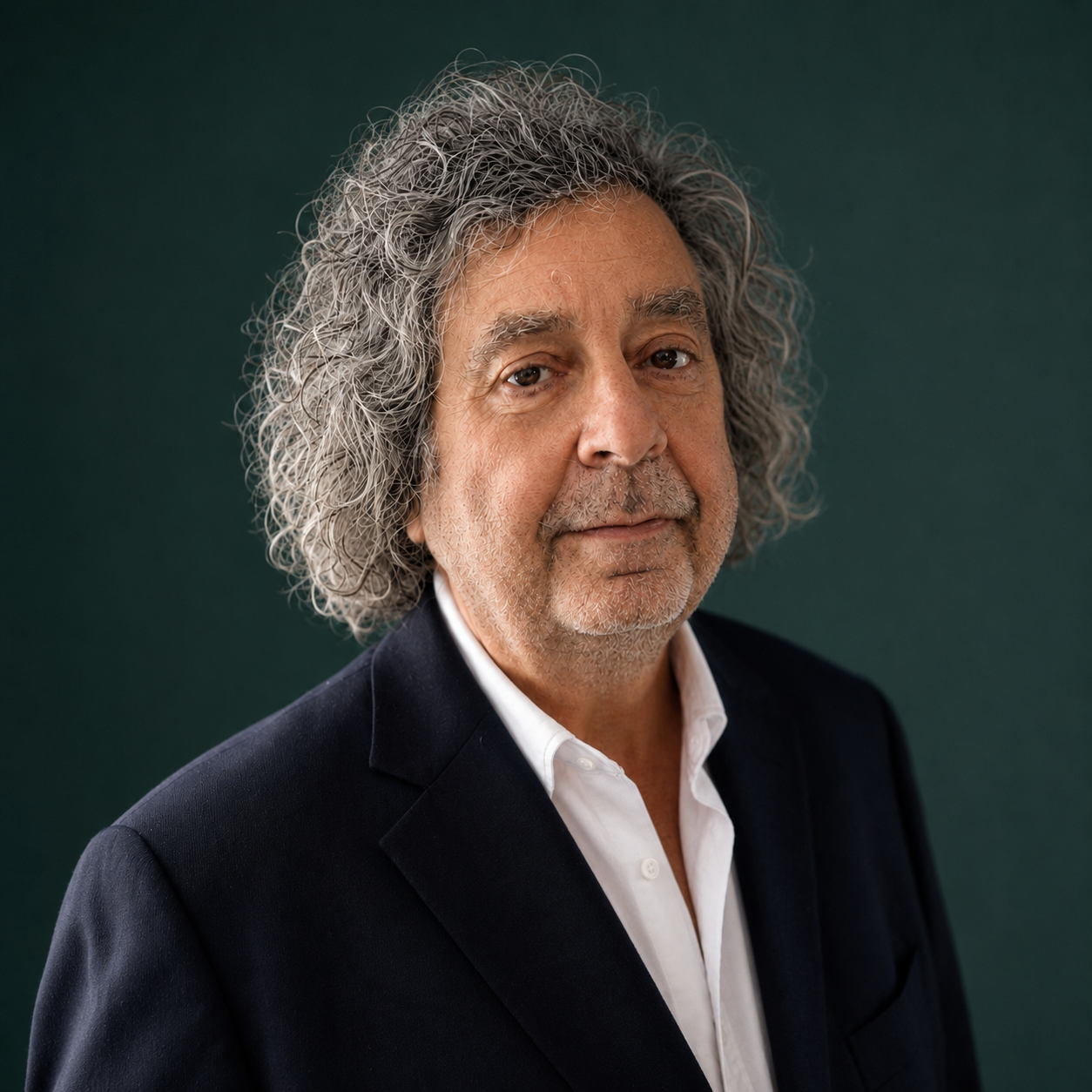 Headshot of a mature man with curly gray hair, wearing a dark suit jacket and white collared shirt, against a dark green background.