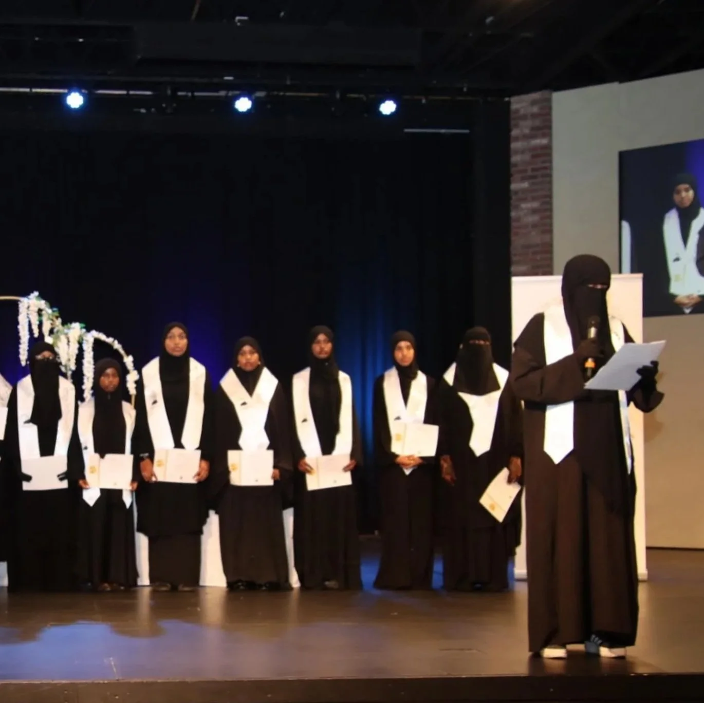 Group of people in graduation gowns and hijabs standing on stage during a graduation ceremony, with one person reading from a paper at a podium, and a large screen displaying a close-up of them.