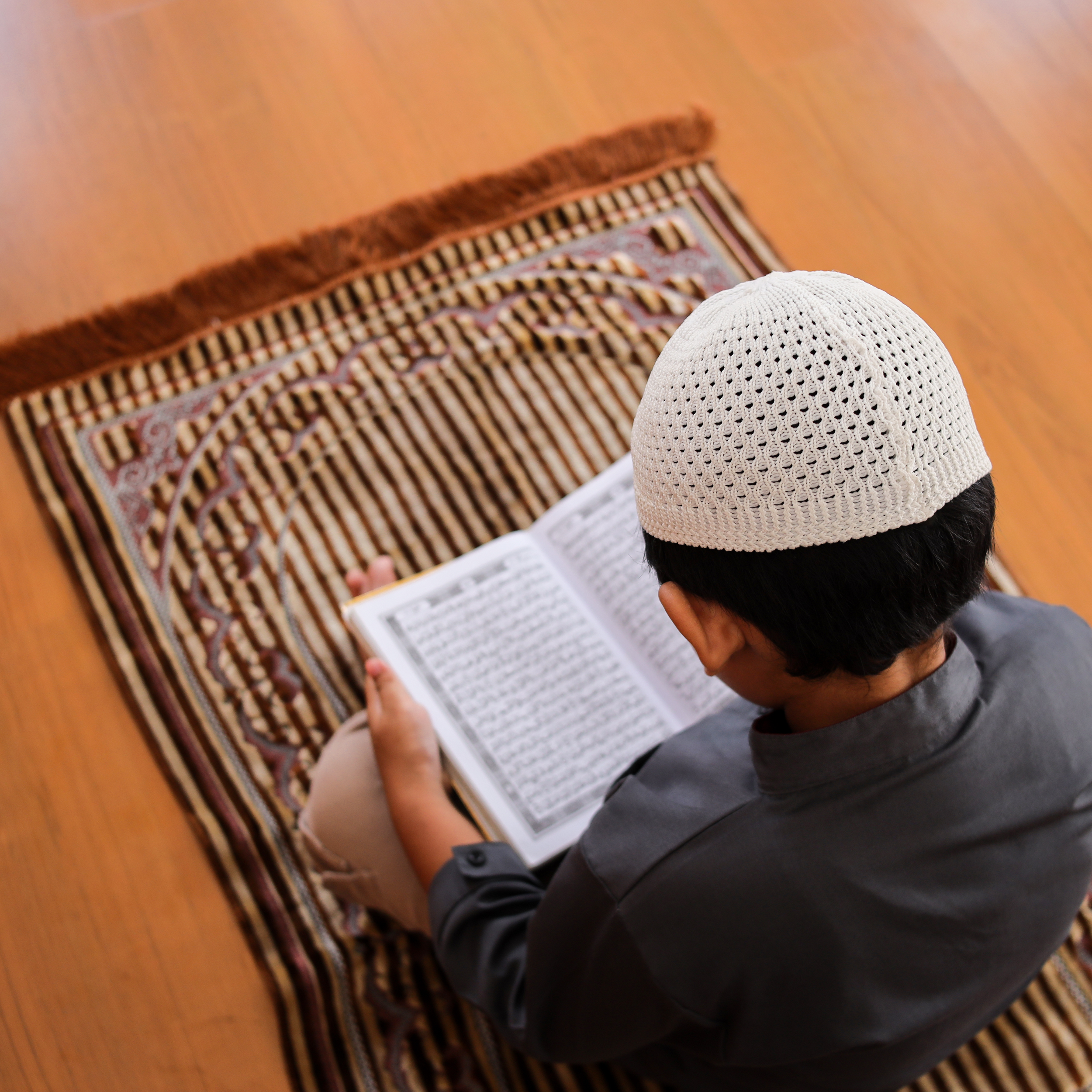 Close-up of a person wearing a white knitted cap, sitting on the floor, reading a book, with a patterned rug and wooden flooring visible.