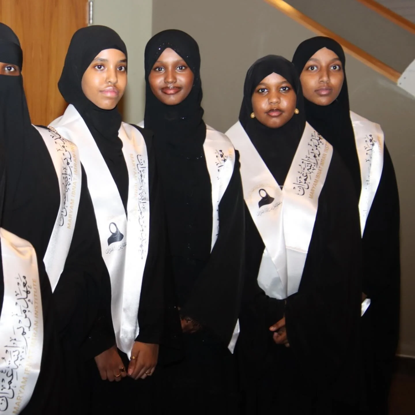 Group of women wearing black hijabs and graduation sashes at an indoor ceremony.