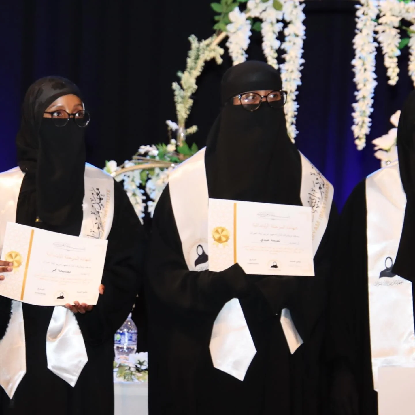 Three women in black abayas and niqabs holding certificates at a graduation ceremony, with floral decorations in the background.