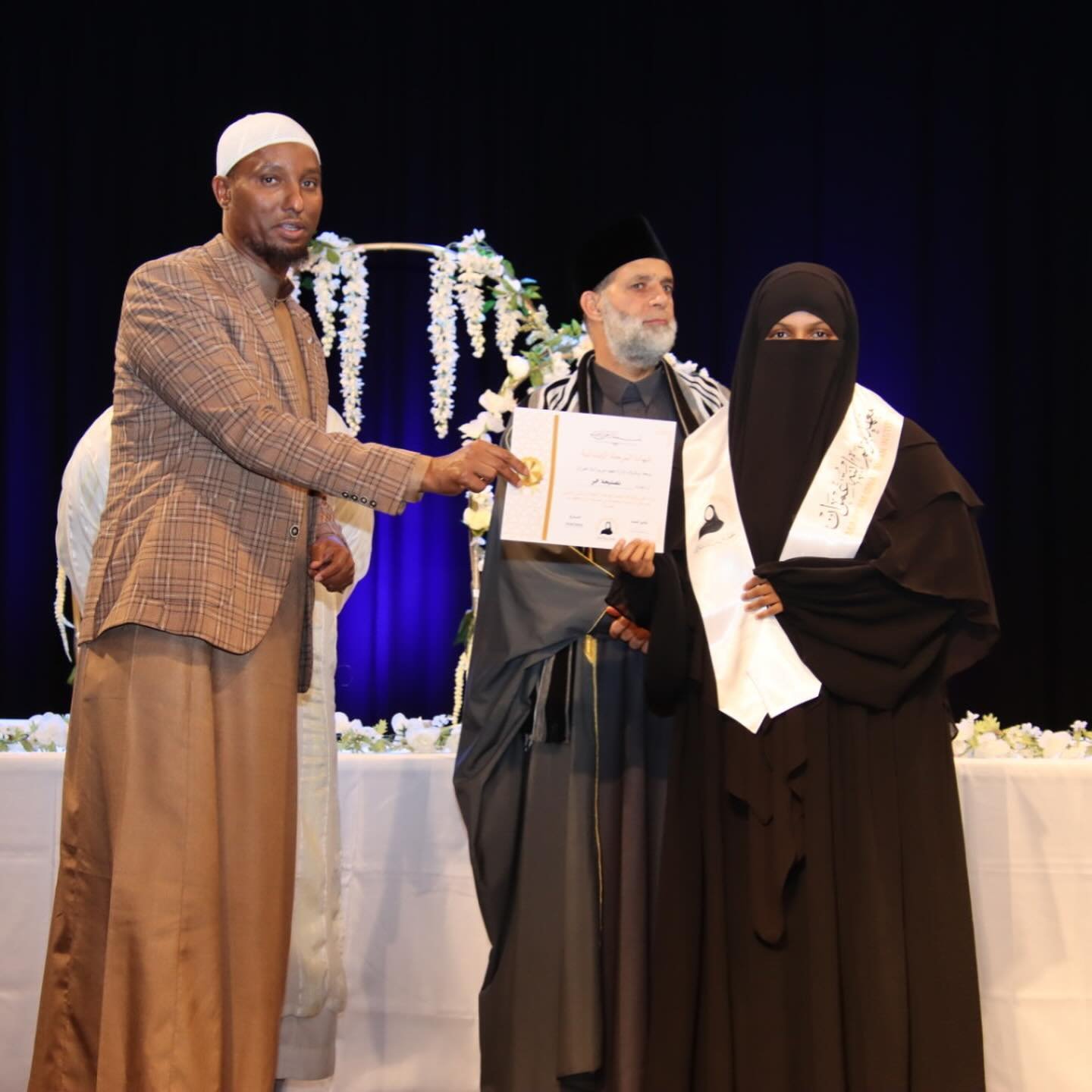 People on stage during a ceremony, a man in religious attire receiving an award from another man, with a woman in Islamic attire standing nearby and holding a certificate.