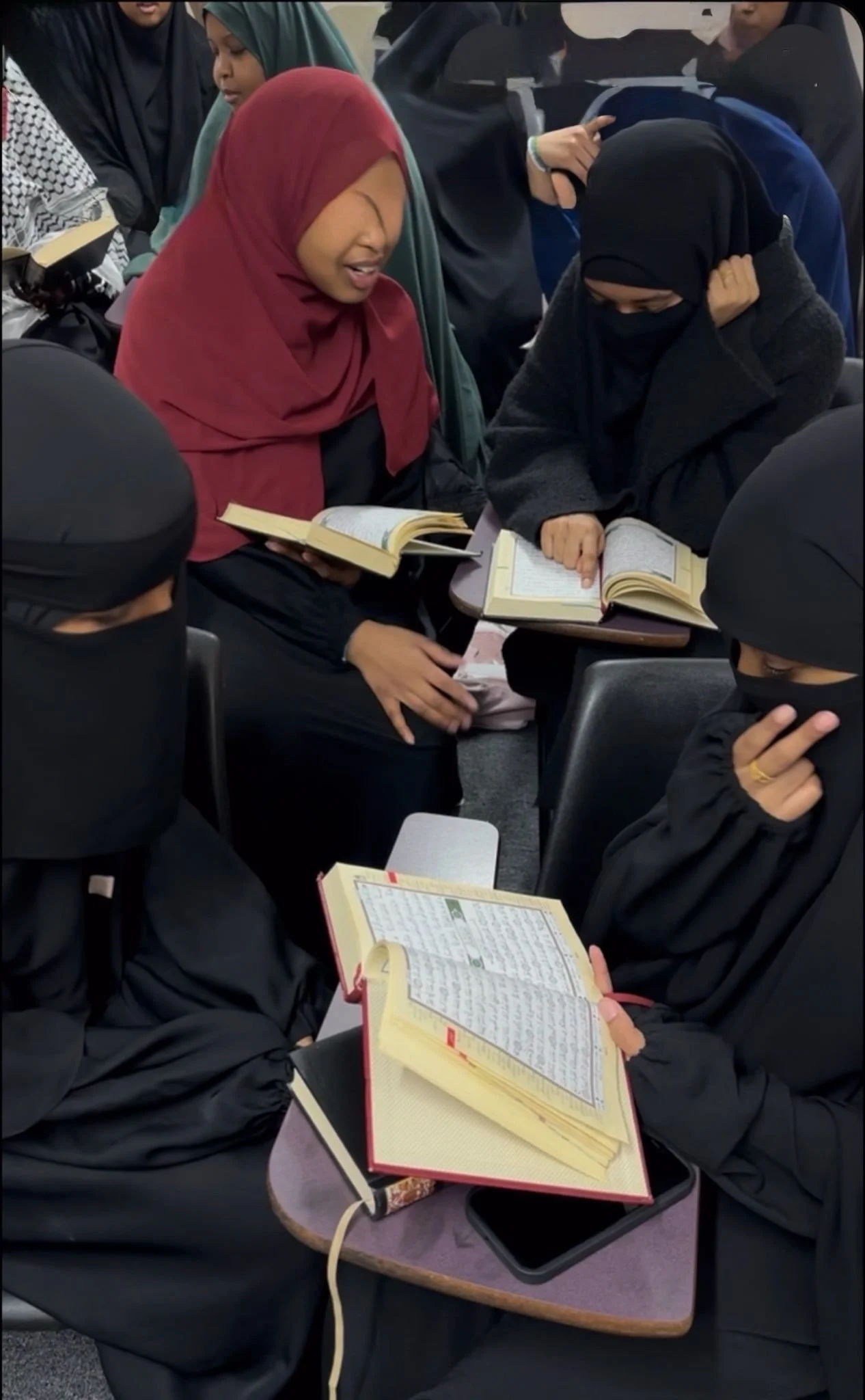 The image shows a person reading a Quran, which is open on their lap. The person is wearing a pink or peach-colored garment. The background includes a colorful patterned carpet and a purple or maroon textured floor.