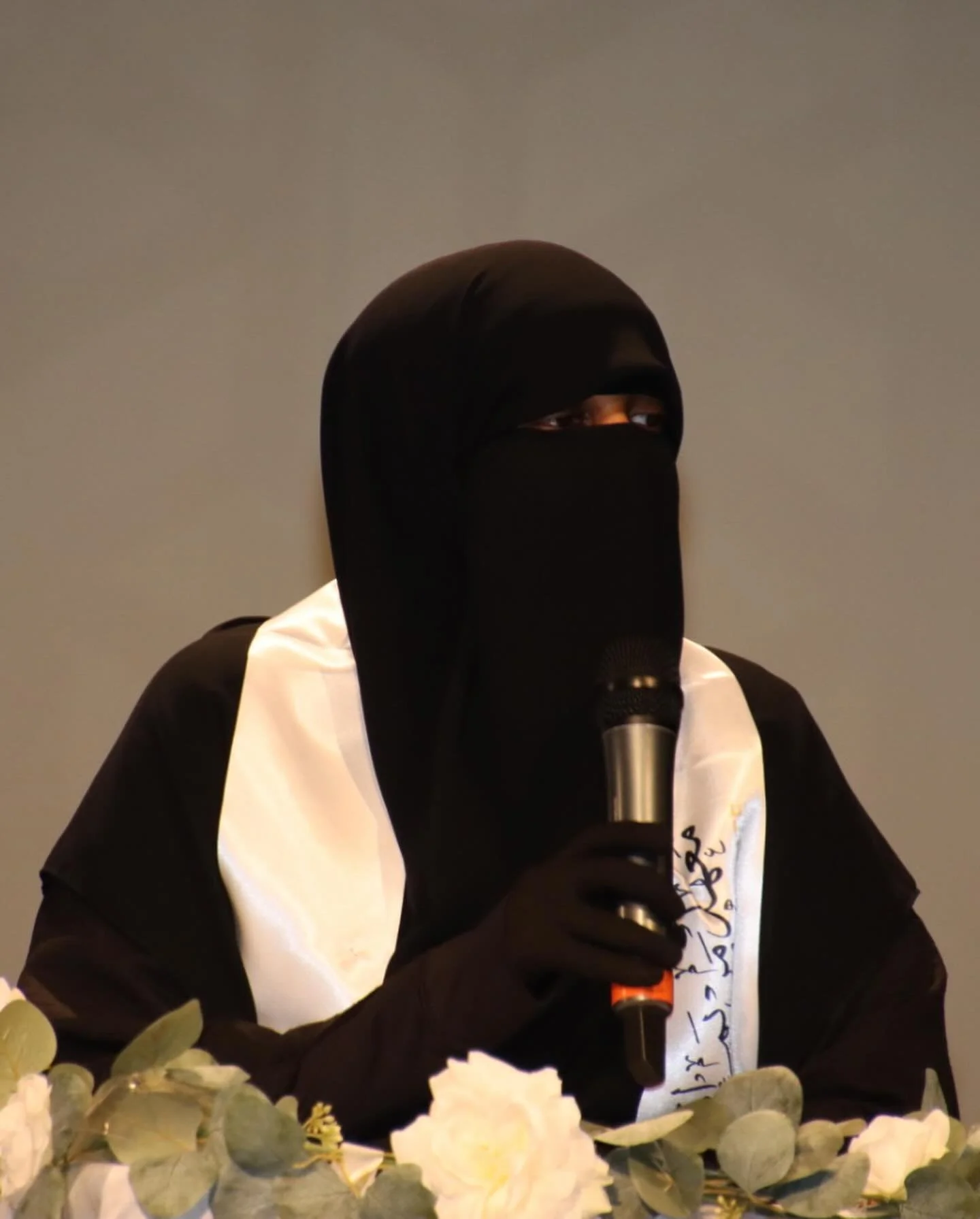 Person wearing a black niqab and a white sash, holding a microphone, surrounded by white flowers.