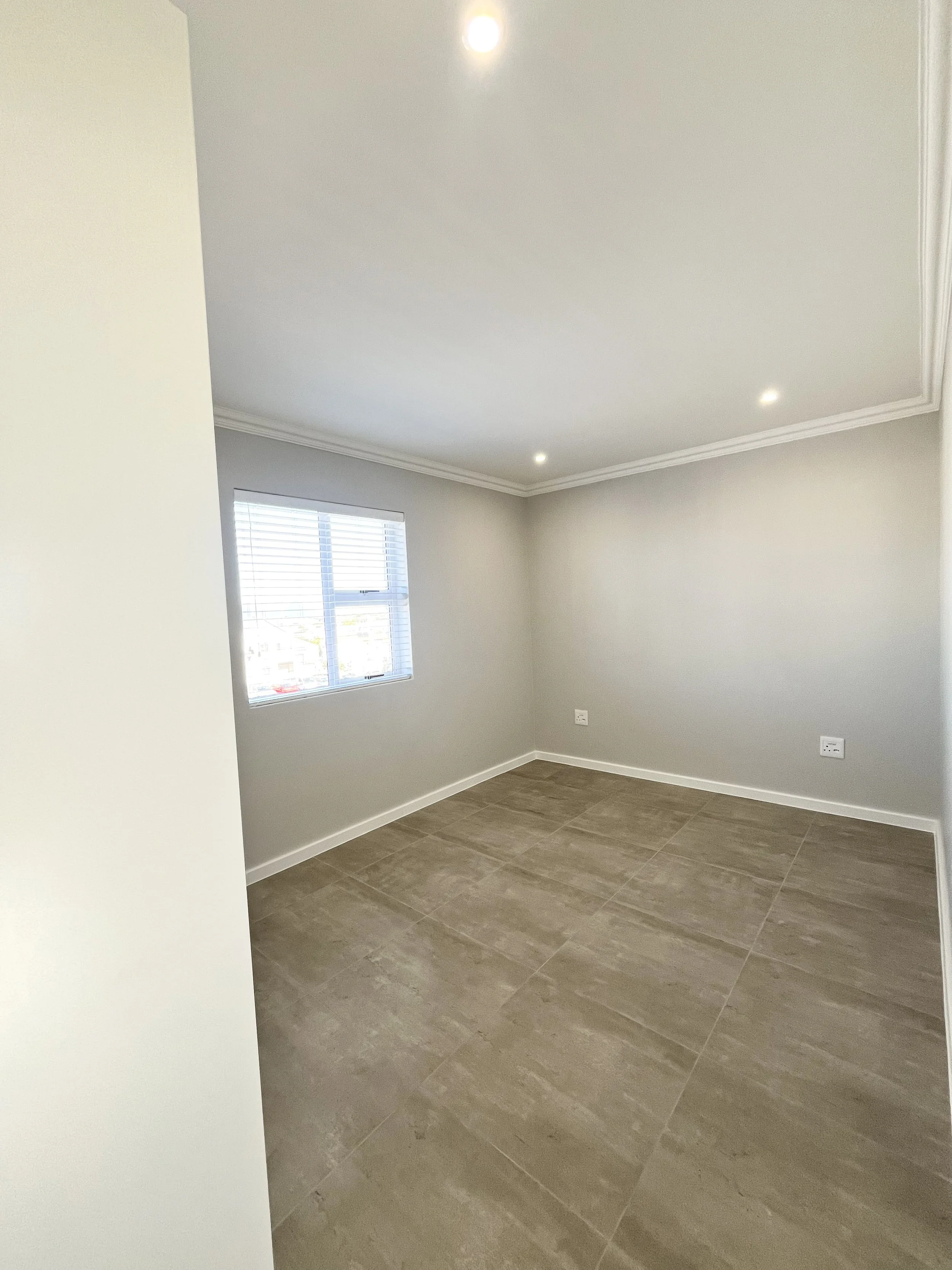Interior of an unfurnished room with gray walls, a window with blinds, beige tiled flooring, white baseboards, and a ceiling with crown molding and recessed lighting.