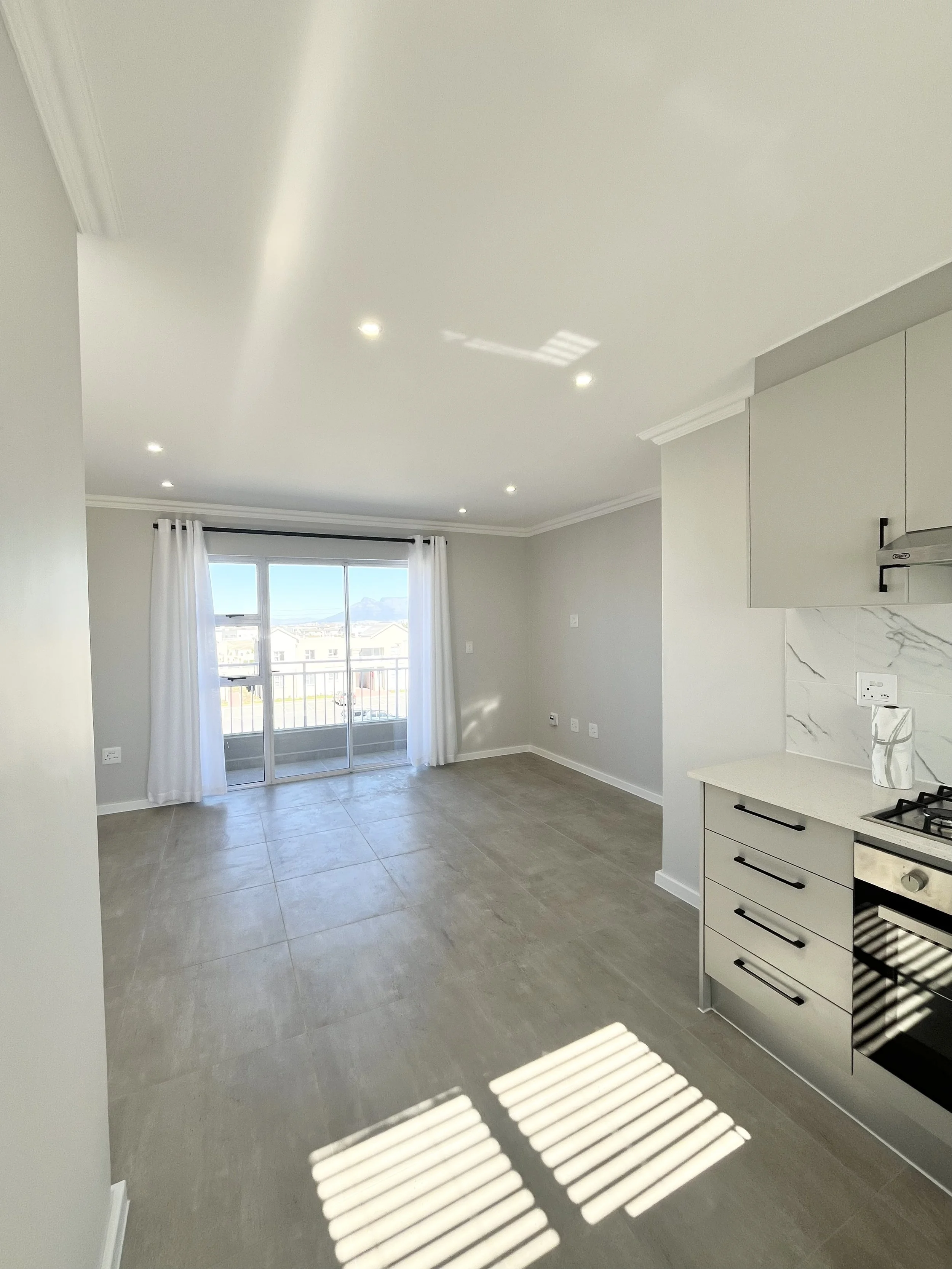 An interior view of a modern, minimally decorated kitchen and living area with large sliding glass doors leading to a balcony, gray tiled floors, white curtains, and some kitchen cabinetry.