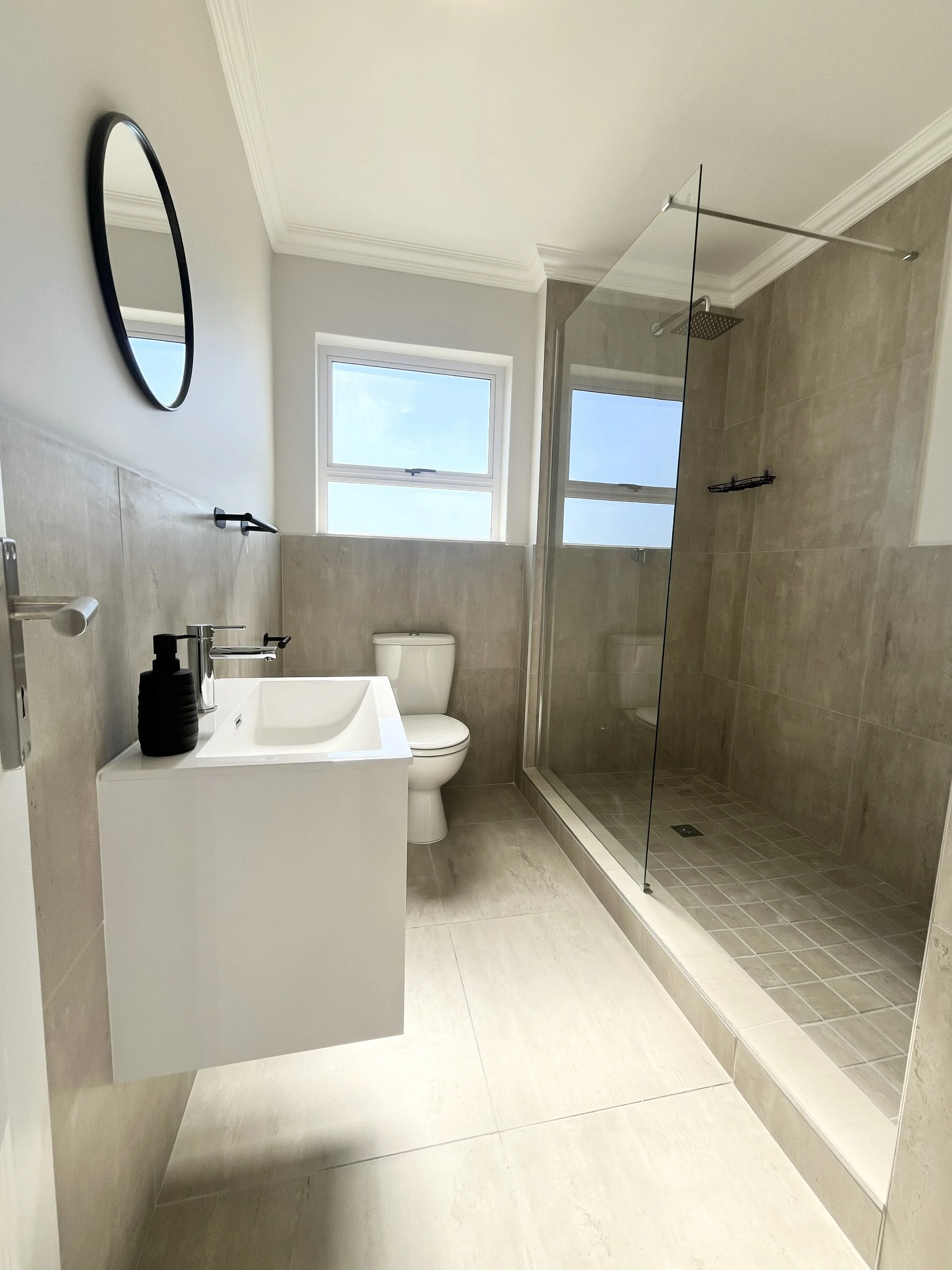 A modern bathroom with a walk-in shower, white sink with black soap dispenser, toilet, and a window with natural light.