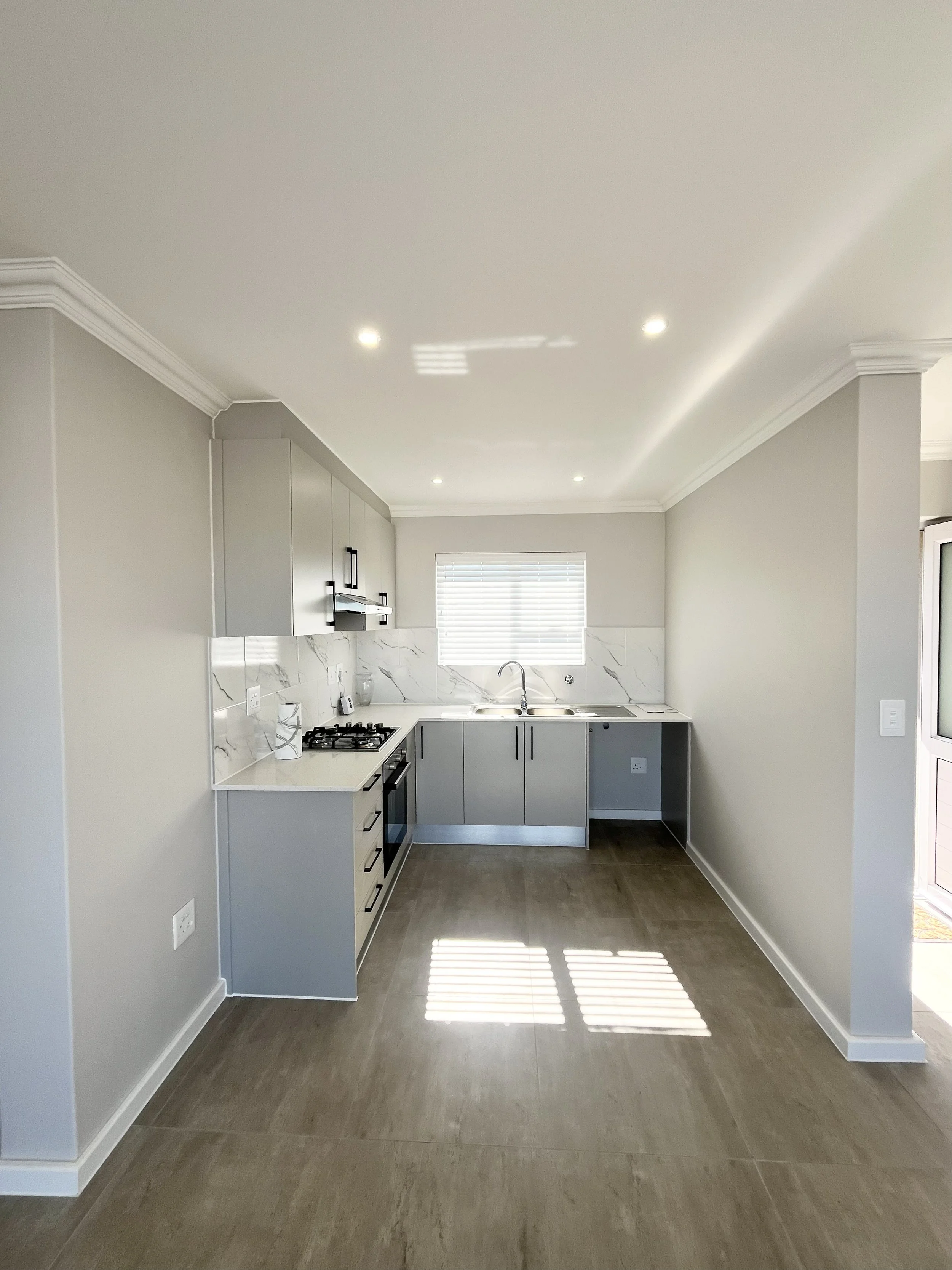 Modern kitchen with gray cabinets, white marble backsplash, window with closed blinds, and brown wood floor with sunlight reflections.