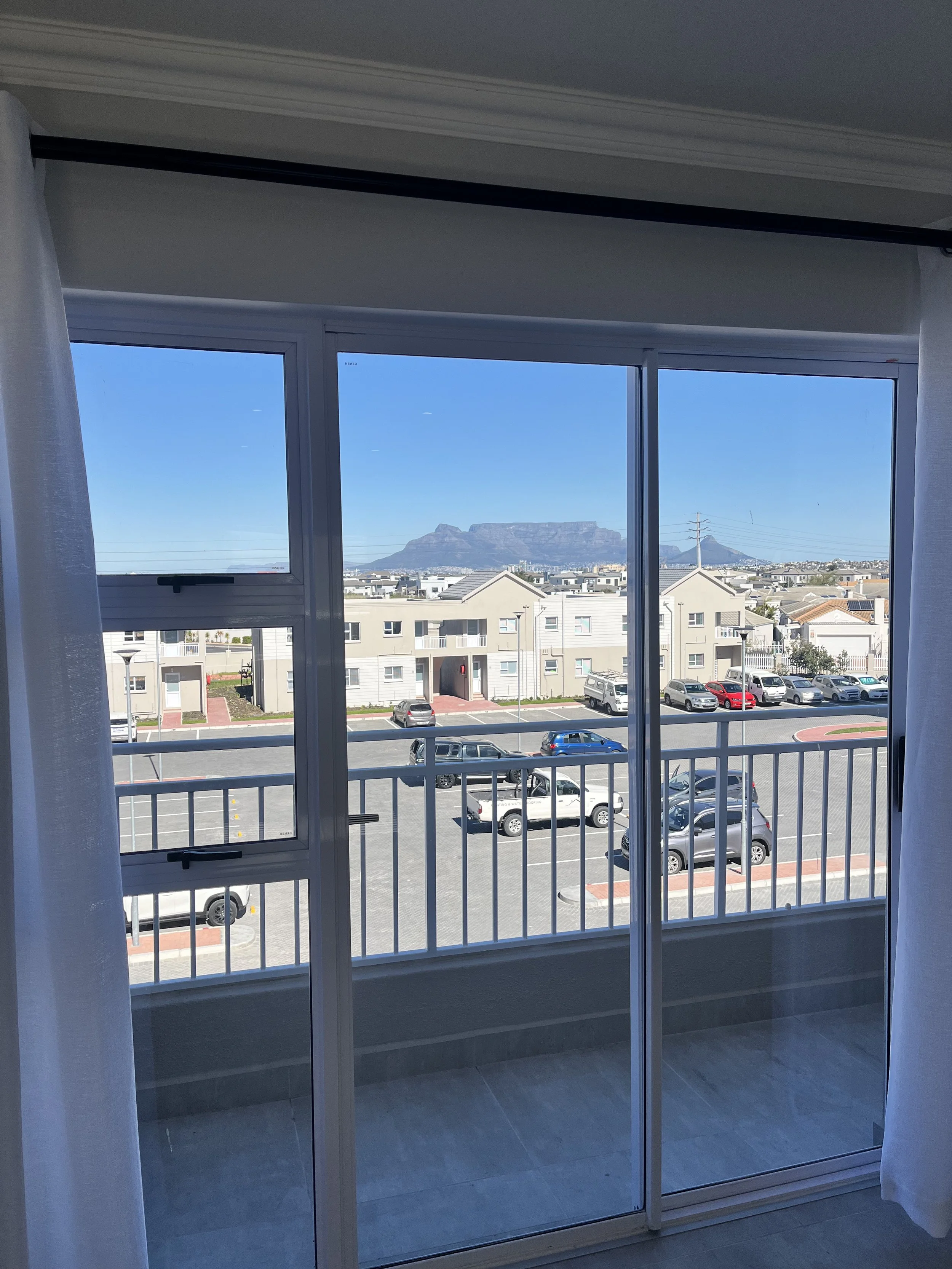 View through glass sliding doors showing a parking lot with several cars, residential buildings, and a mountain in the distance under a clear blue sky.