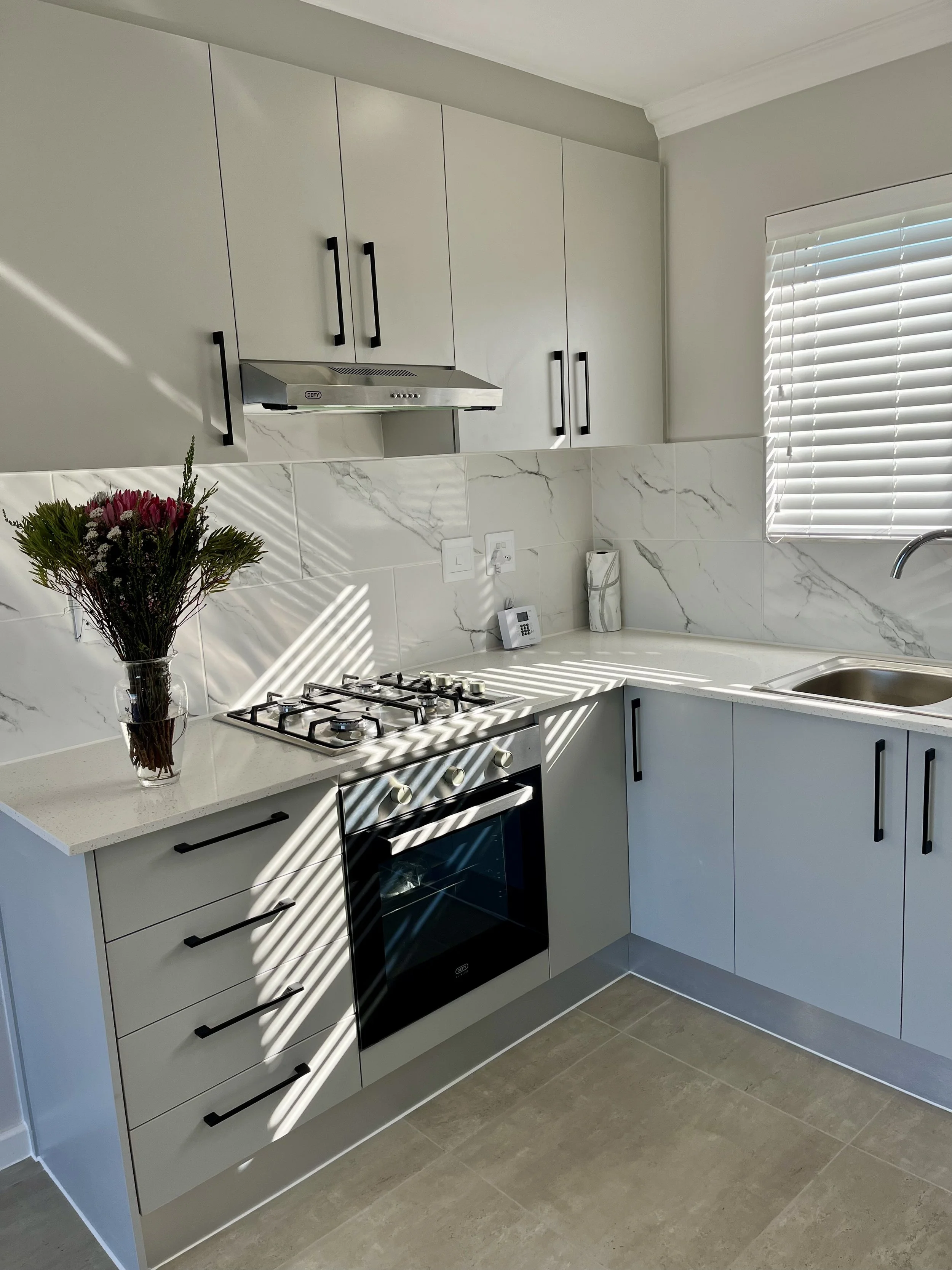 A modern kitchen with white cabinets, a marble backsplash, and a window with white blinds. There's a vase with flowers on the counter, a gas stove, and a range hood.