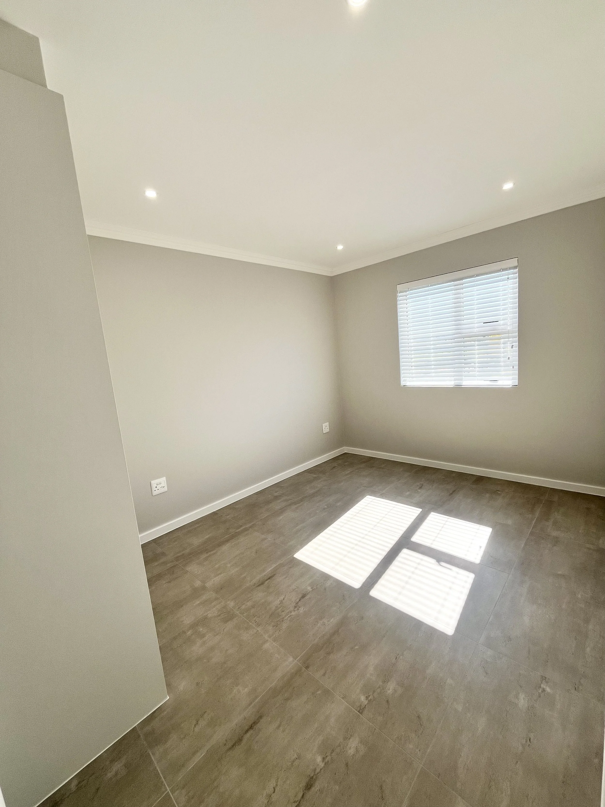 An empty room with beige walls, a window with blinds, and brown tiled flooring, with sunlight casting shadows through the blinds.