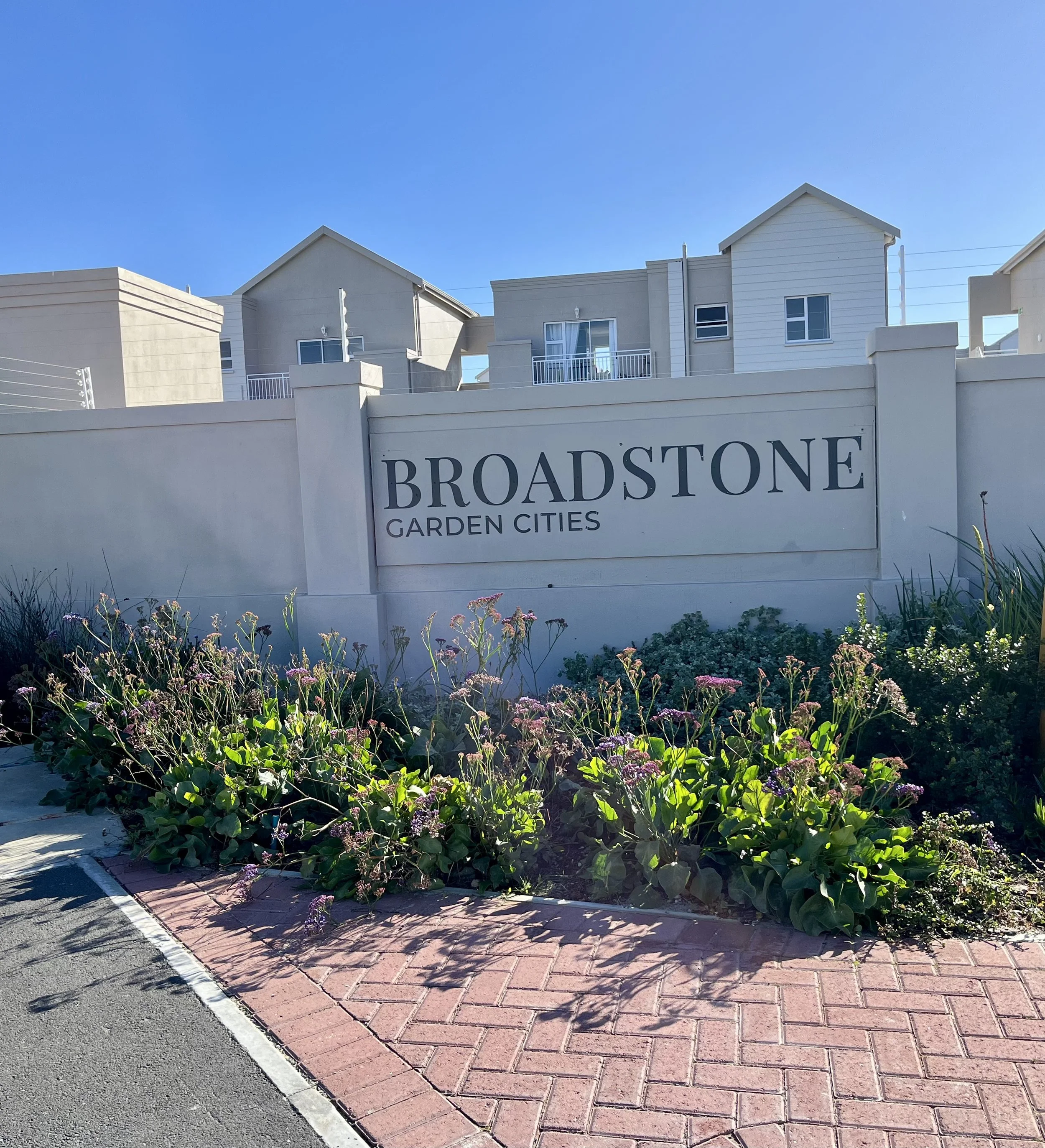 A sidewalk and brick-paved area with a flower bed of green leaves and purple flowers in front of white residential buildings. A sign reading 'Broadstone Garden Cities' is visible on the wall.