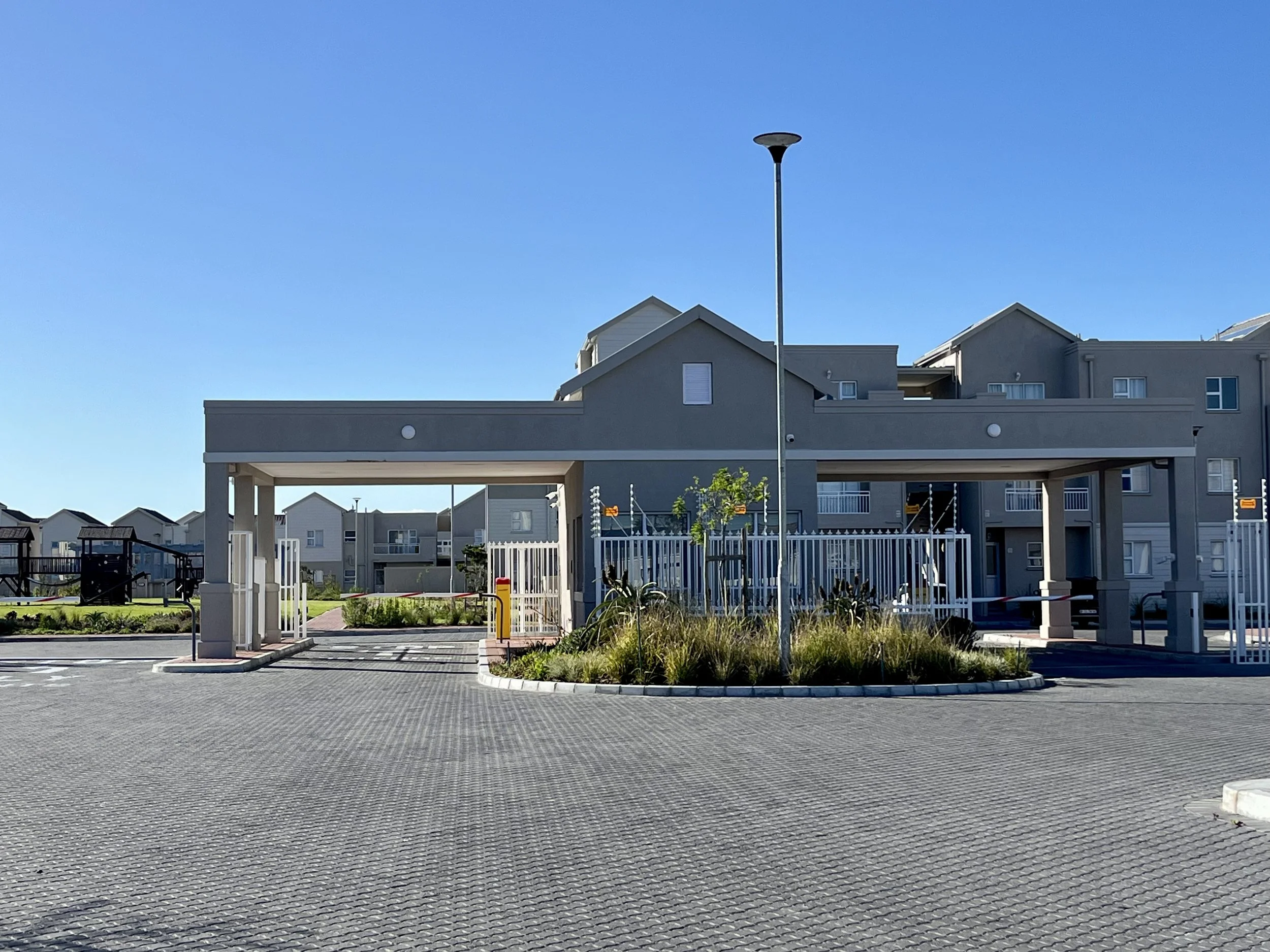 A residential complex with multi-story buildings, a parking area, a gated entrance, and landscaping, under a clear blue sky.