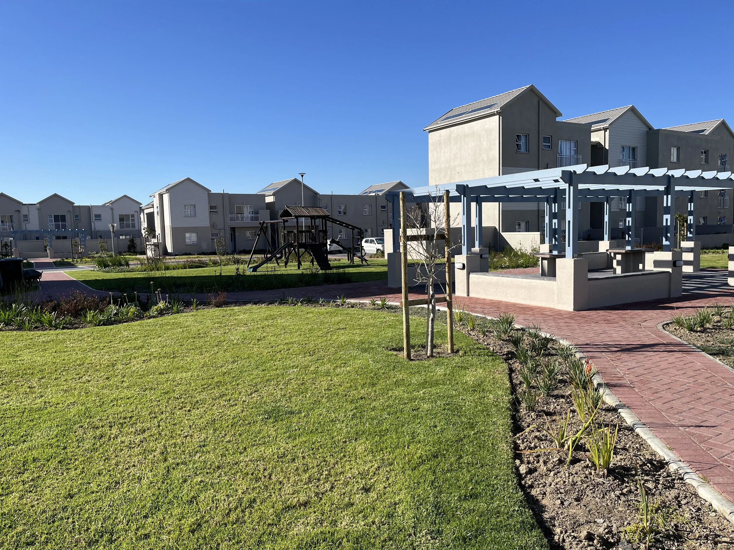A residential complex with multiple multi-story townhouses, a playground, a pergola, and a landscaped garden with small trees and plants.