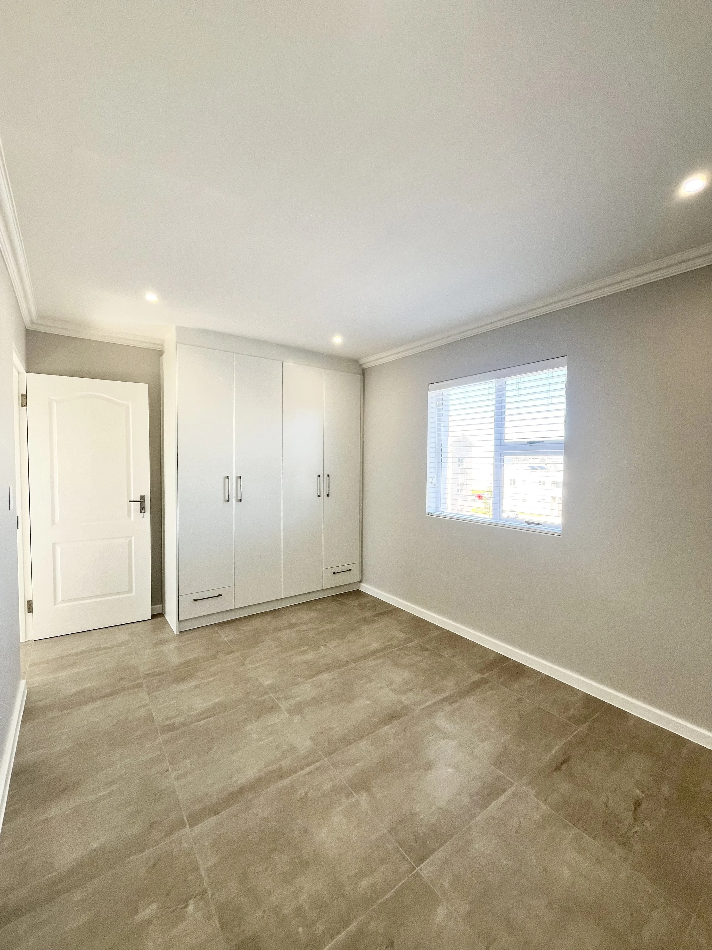 A room with beige tiled flooring, gray walls, a white door, a window with blinds, and a white built-in wardrobe with silver handles.