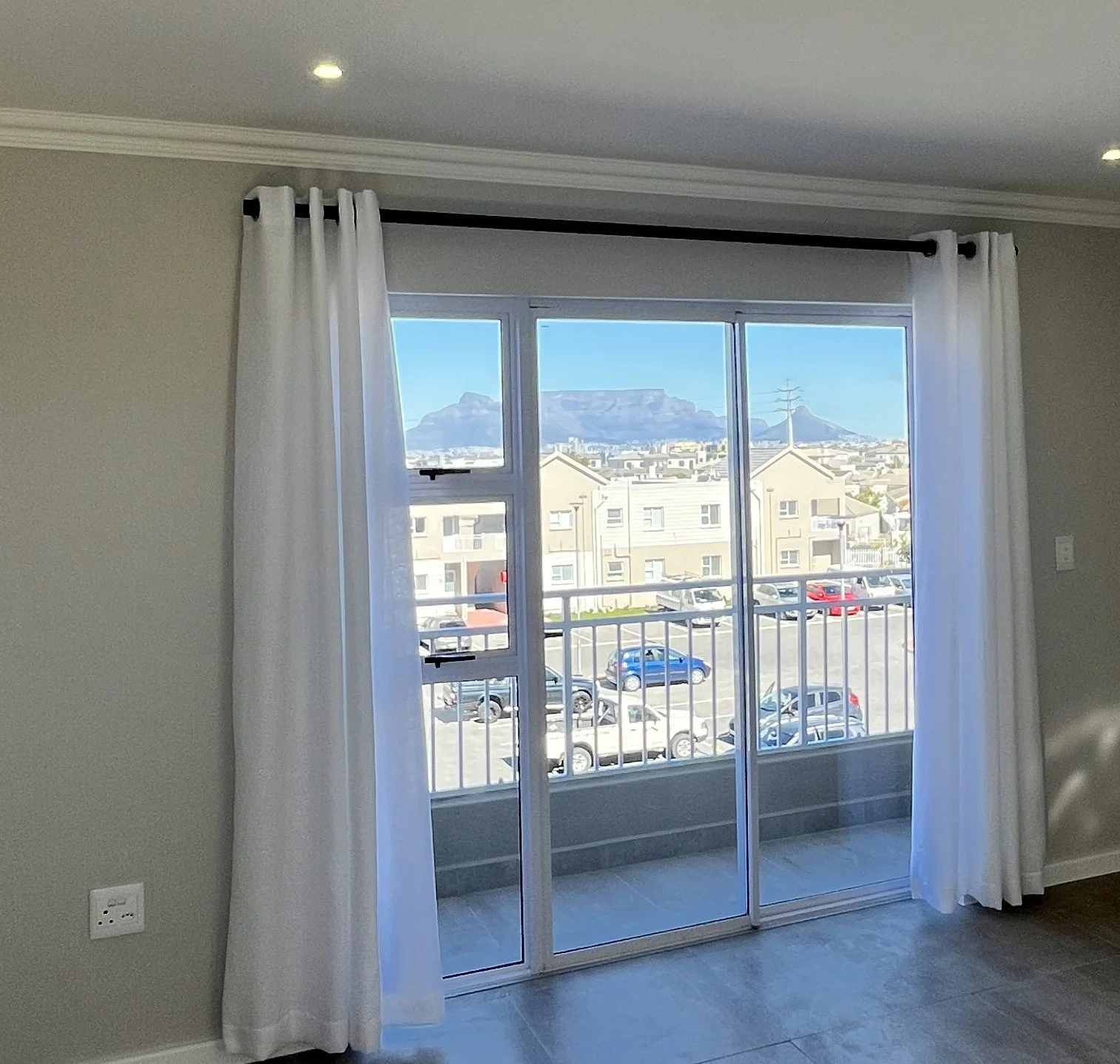 Interior view of a room with a sliding glass door leading to a balcony, showing a cityscape and mountains in the distance through the window, with white curtains on a black curtain rod.