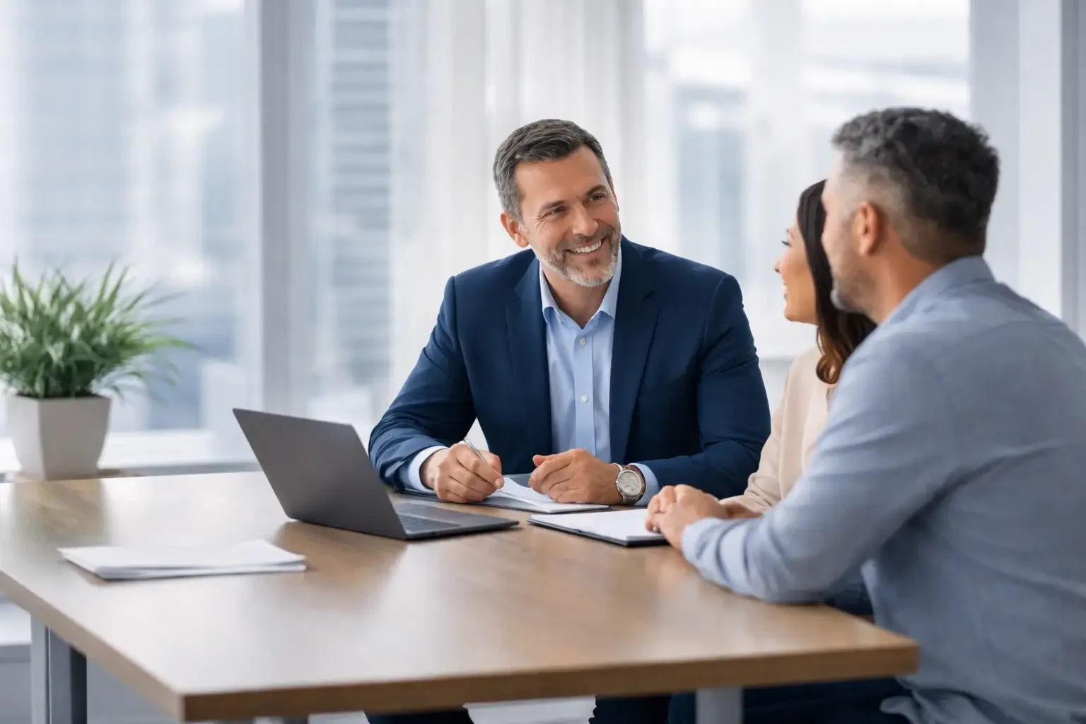 Three professionals having a meeting at a conference room table, smiling and engaging in conversation, with a laptop and documents on the table.