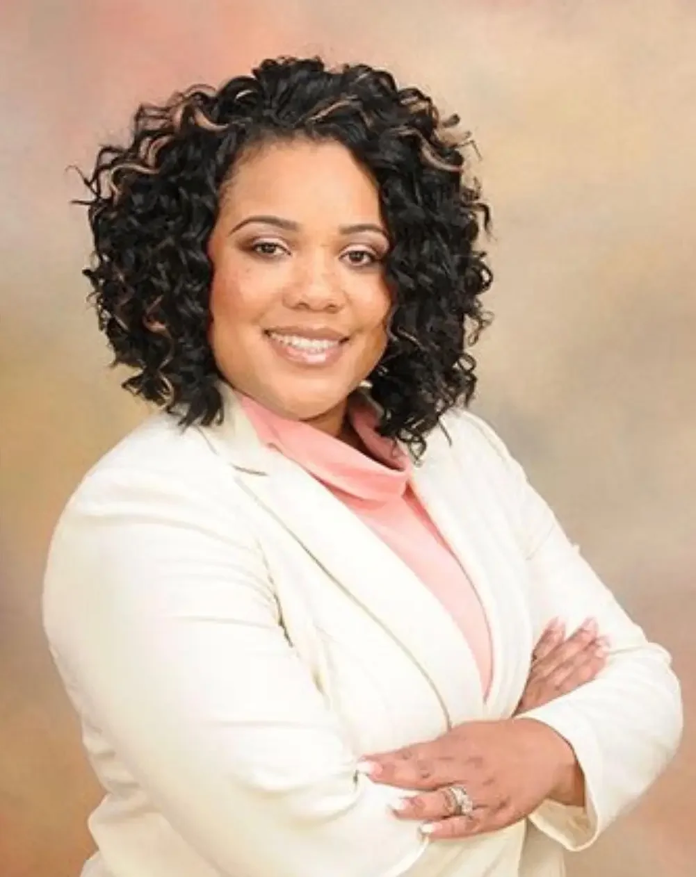 A woman with curly black hair, crossed arms, wearing a white blazer and light pink top, smiling against a beige background.