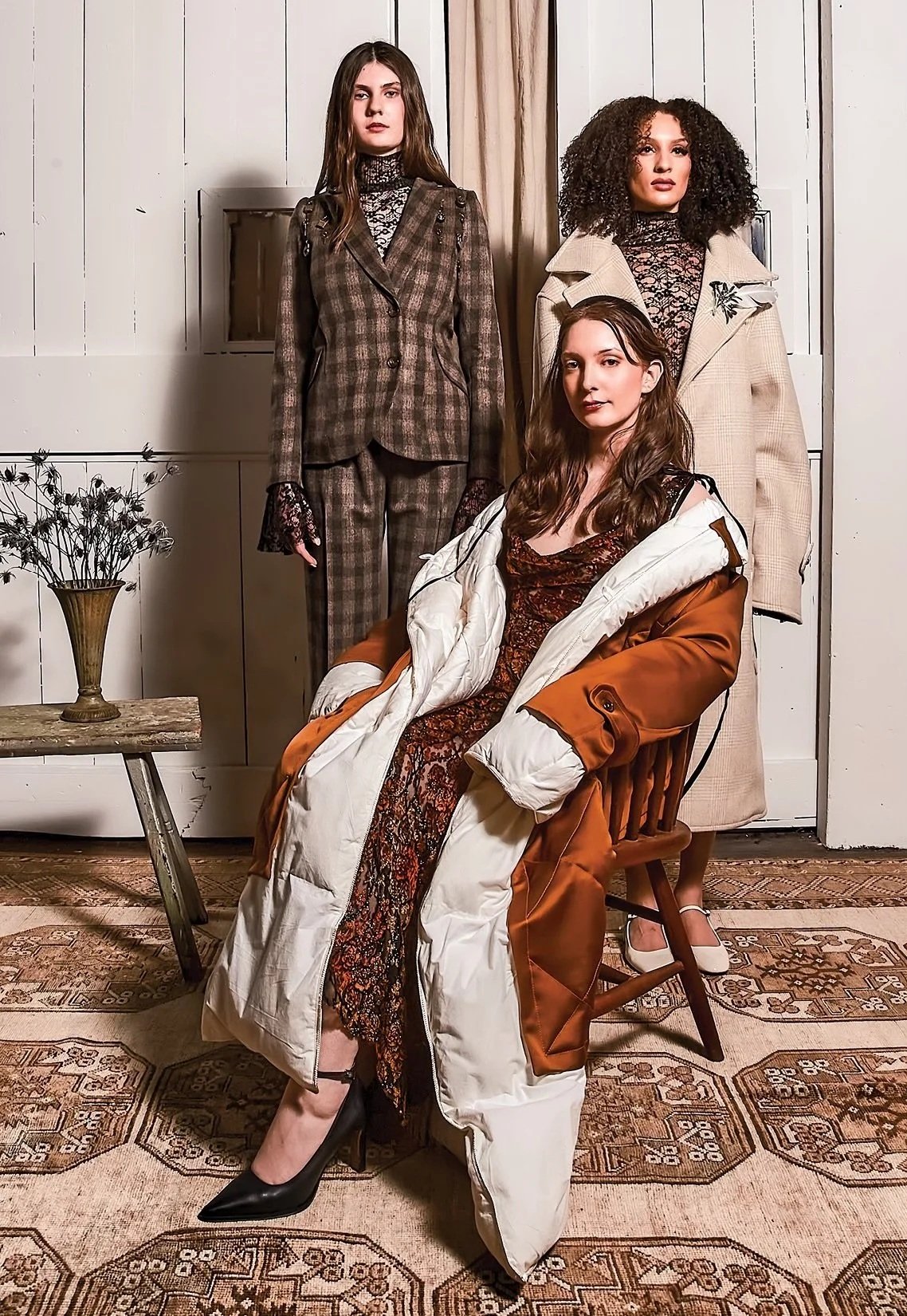 Three women in fashionable outfits pose indoors against a white paneled wall with a vase of dried flowers on a small wooden table and a patterned rug on the floor. David Ferron fashion design background from Parsons School of Design.