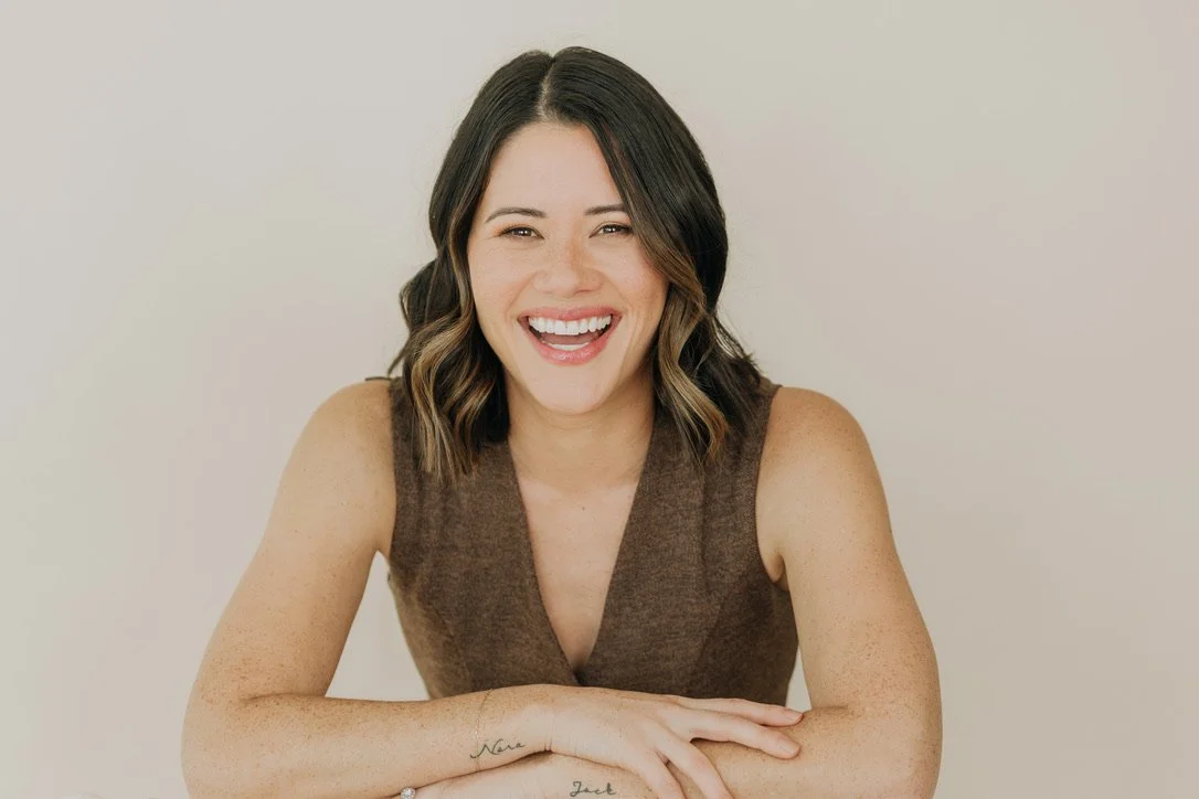 A woman with shoulder-length wavy dark hair with highlights, smiling and sitting with arms crossed, wearing a sleeveless brown top against a plain light background.
