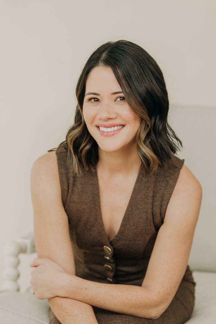 A woman with shoulder-length dark hair with light brown highlights, smiling while sitting on a light-colored couch, wearing a sleeveless brown dress with buttons.