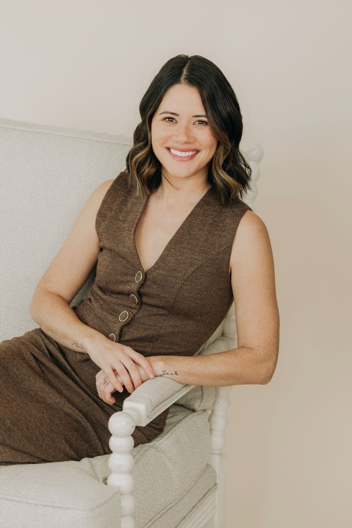 A woman with shoulder-length dark hair styled in loose waves, smiling, seated on a beige armchair against a plain wall, wearing a brown sleeveless dress with buttons down the front.