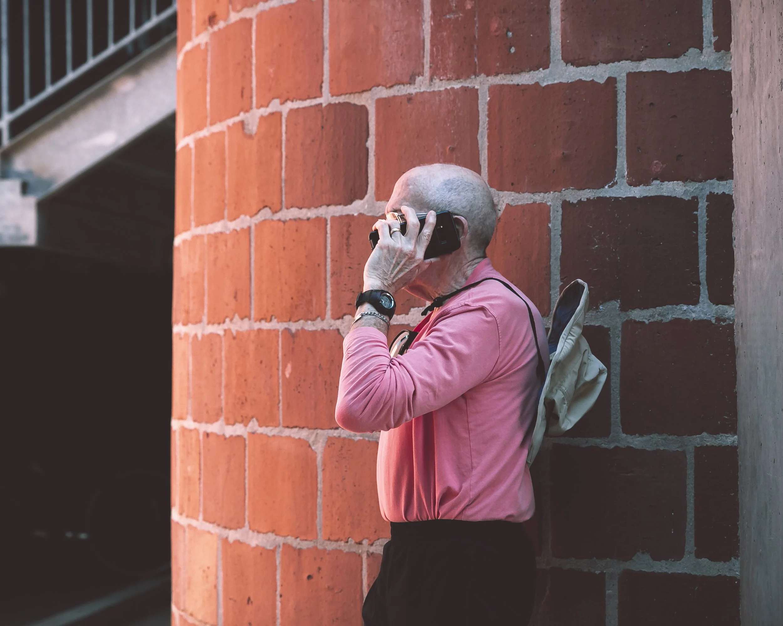 Person with gray hair wearing a pink jacket, holding a camera, leaning against a brick wall.