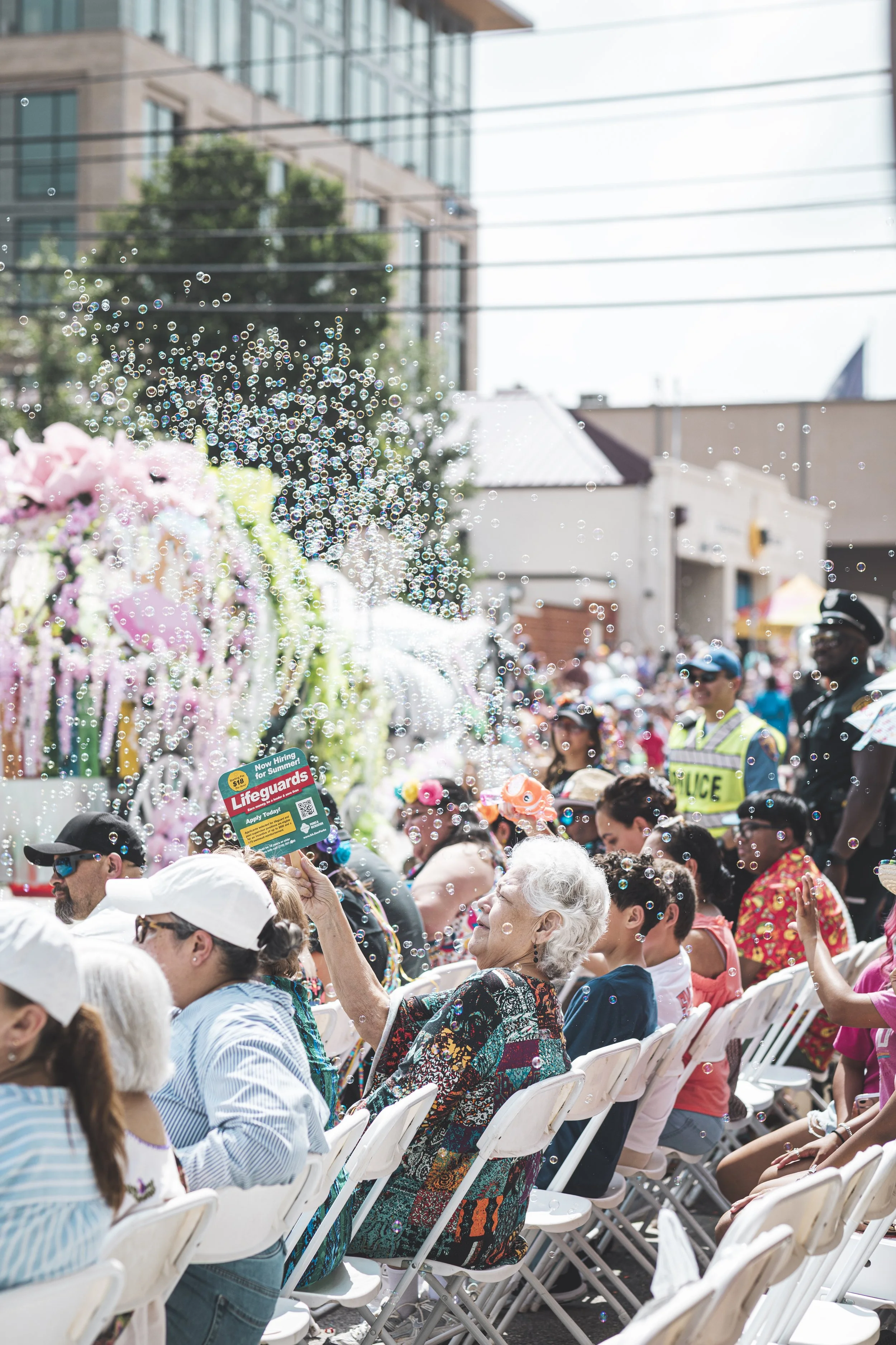 A crowded outdoor event with many people sitting on white chairs, some of them smiling and taking photos. Bubbles are floating in the air, and there are buildings and trees in the background. One woman is holding a colorful flyer that says "Lifeguard