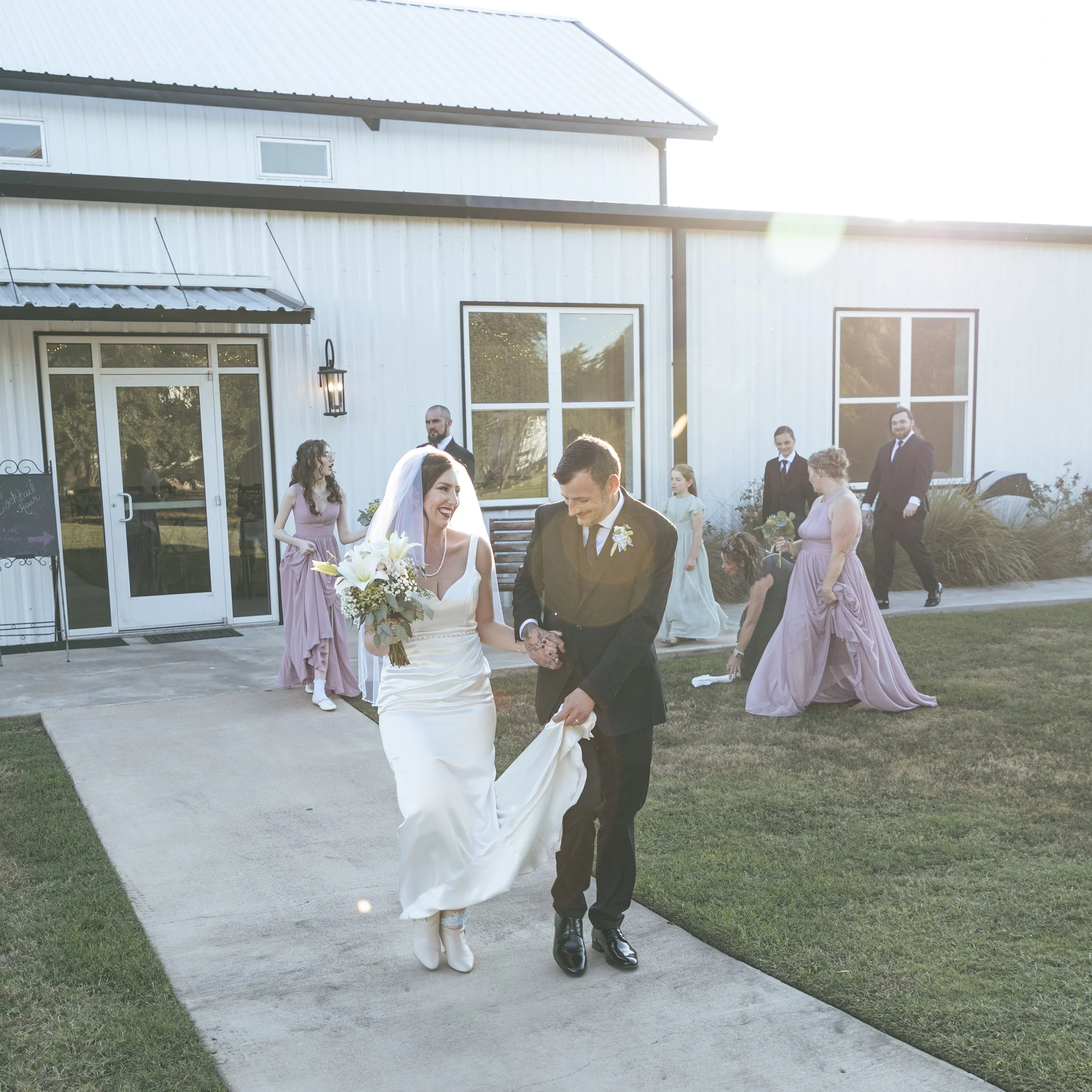 A wedding ceremony outside a white building with large windows and trees reflected in the glass. The bride and groom are holding hands, smiling, surrounded by bridesmaids and groomsmen. The bride wears a white dress and veil, holding a bouquet of whi