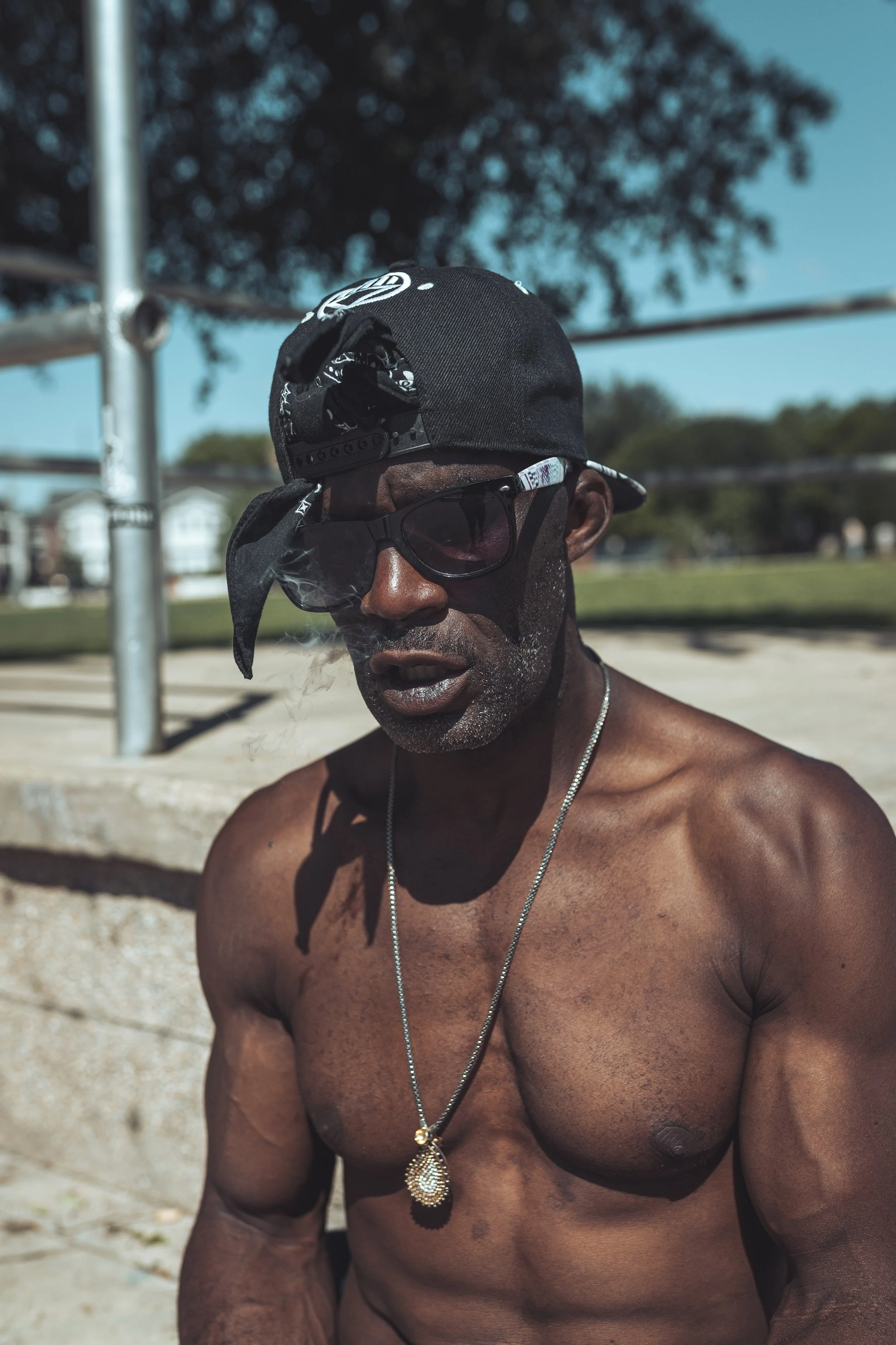 A shirtless man with dark skin, wearing sunglasses, a black cap backward, and a gold necklace with a pendant, standing outdoors near metal bars on a sunny day.