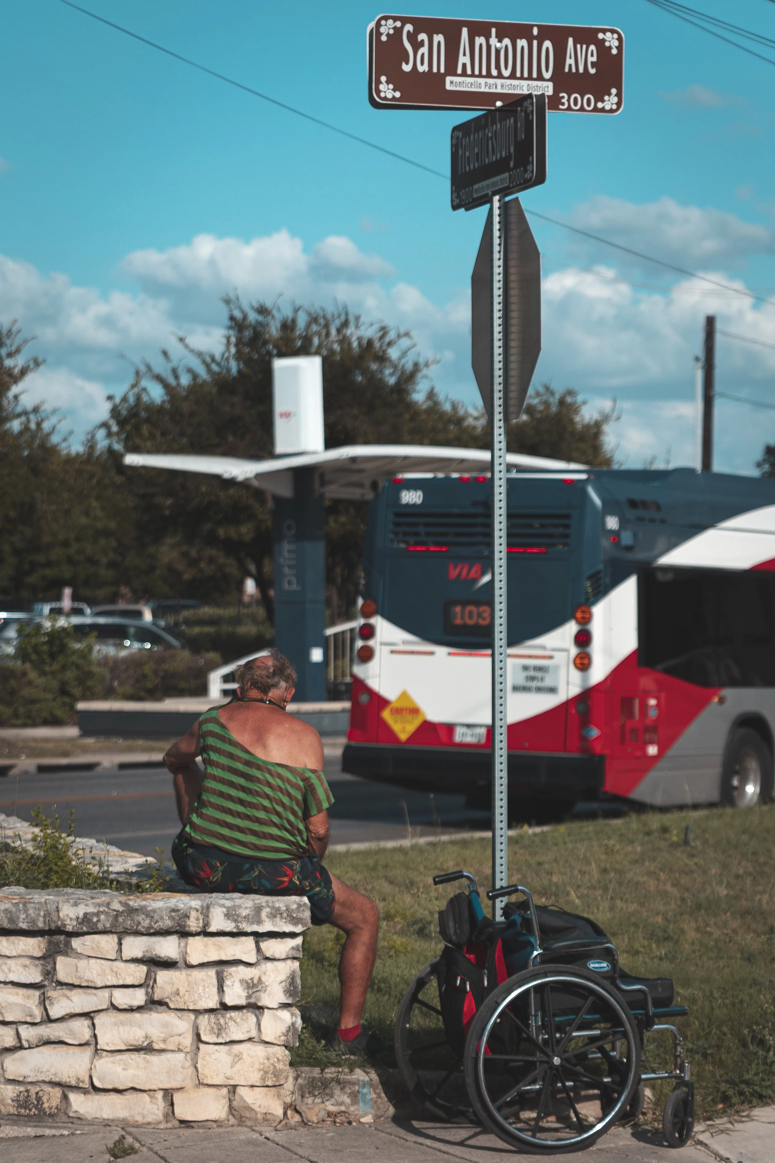 A man in a green and brown striped shirt and colorful shorts is sitting on a stone ledge, with a wheelchair and a black and red backpack and a stroller close by. In the background, there is a red and white bus, trees, and a blue sky with clouds. Ther