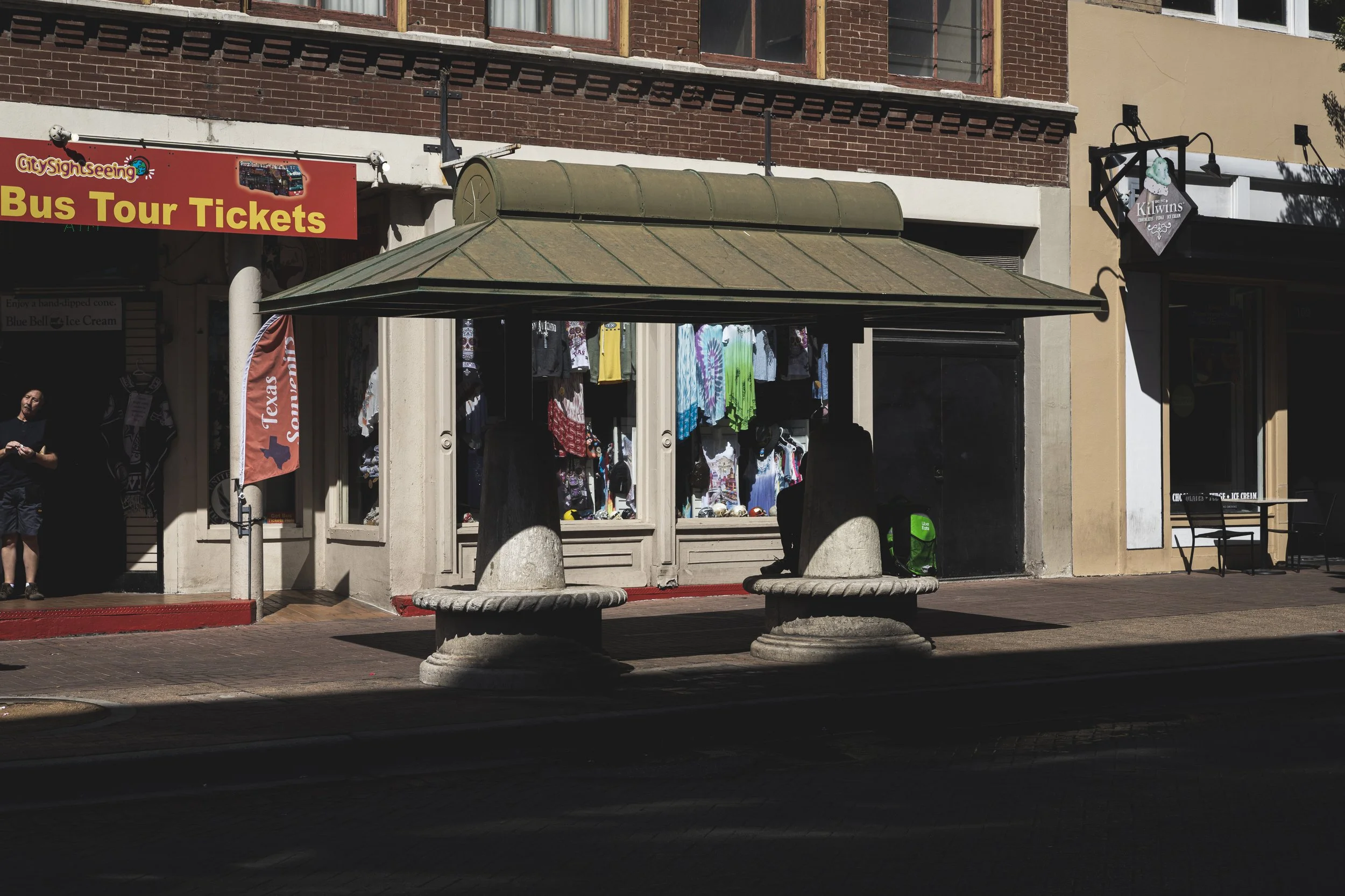 A sidewalk scene featuring a city storefront with clothing displays, a bench with stone bases, and a sign for City Sightseeing Bus Tour Tickets.