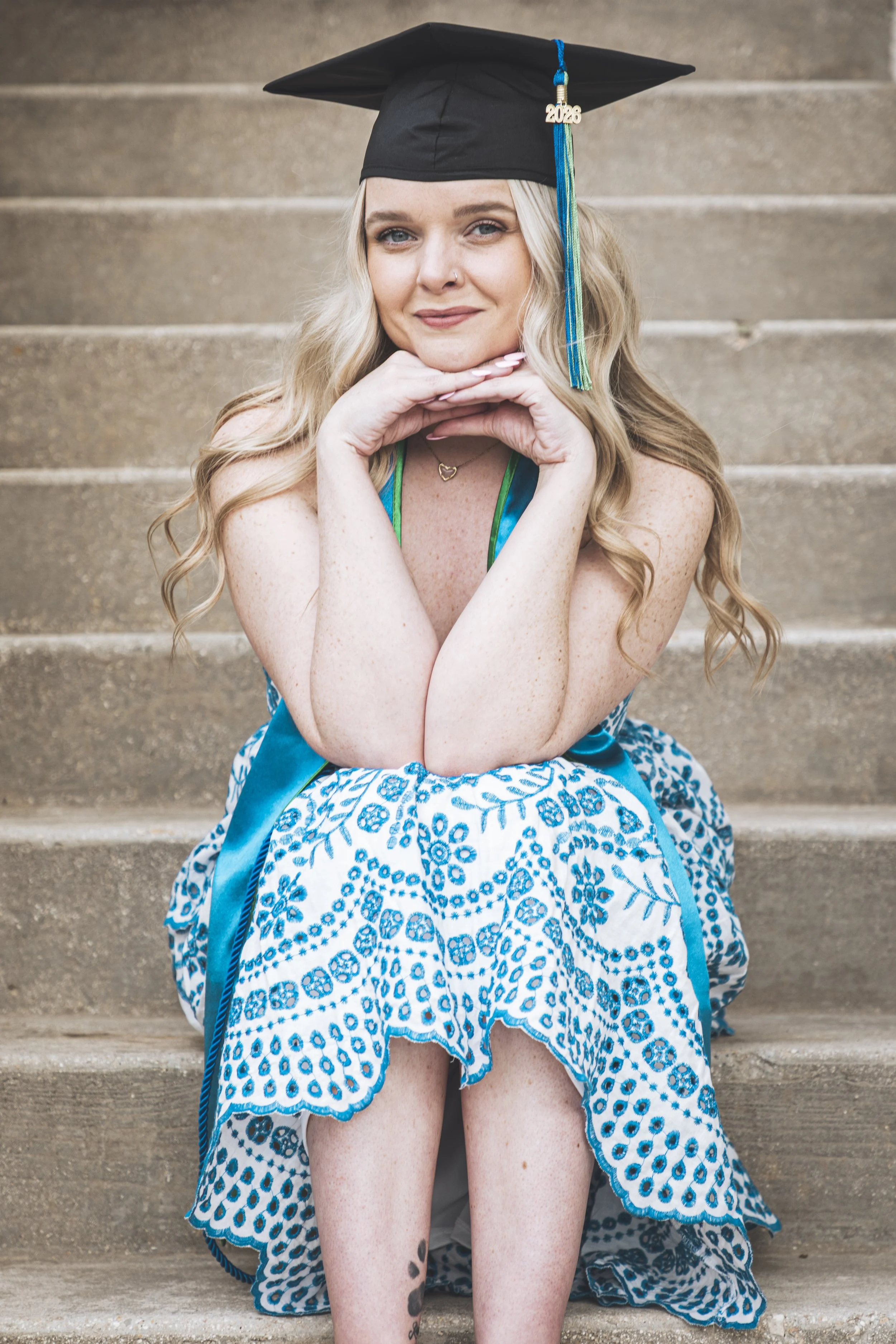 Woman wearing a black graduation cap with a blue and multicolored tassel reading '2028,' sitting on stairs in a blue and white patterned dress, smiling, with her chin resting on folded hands.