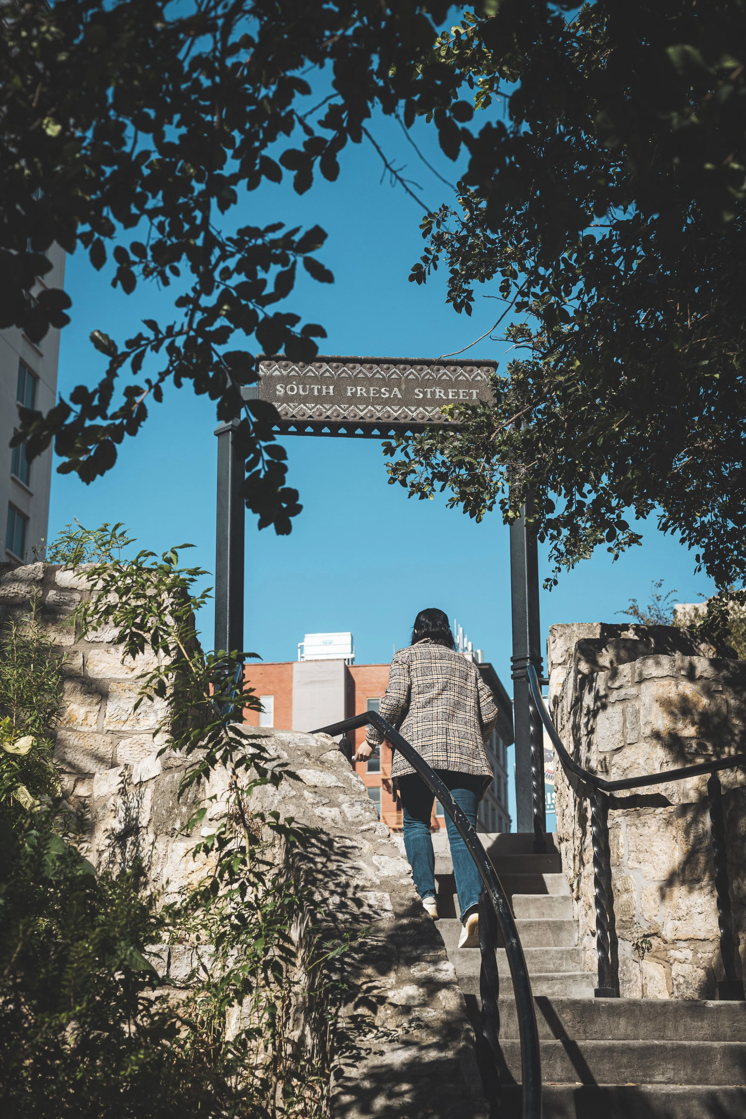 Person ascending stone stairs leading to South Presa Street, framed by trees and a street sign, with a blue sky in the background.
